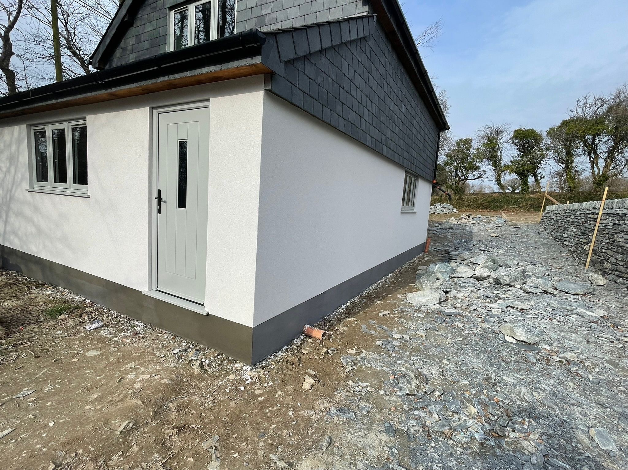 Wykończona elewacja budynku z szarą stolarką okienną i drzwiami, widoczny cokół. W tle kamienny murek i drzewa. Teren wokół budynku w trakcie prac wykończeniowych.