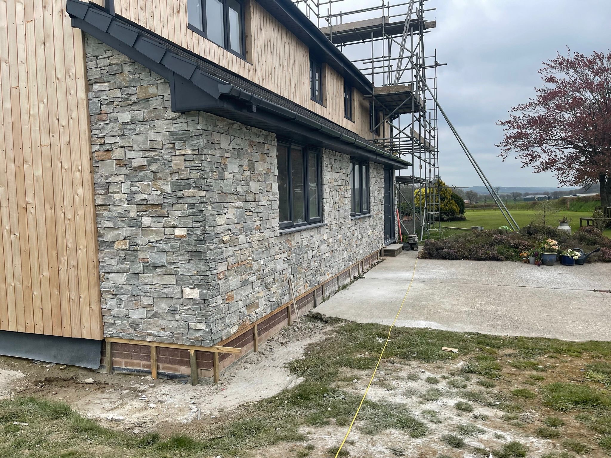 Dom z elewacją z kamienia i drewnianą boazerią, widoczne rusztowania. Detal wykończenia, połączenie materiałów, nowoczesny design.