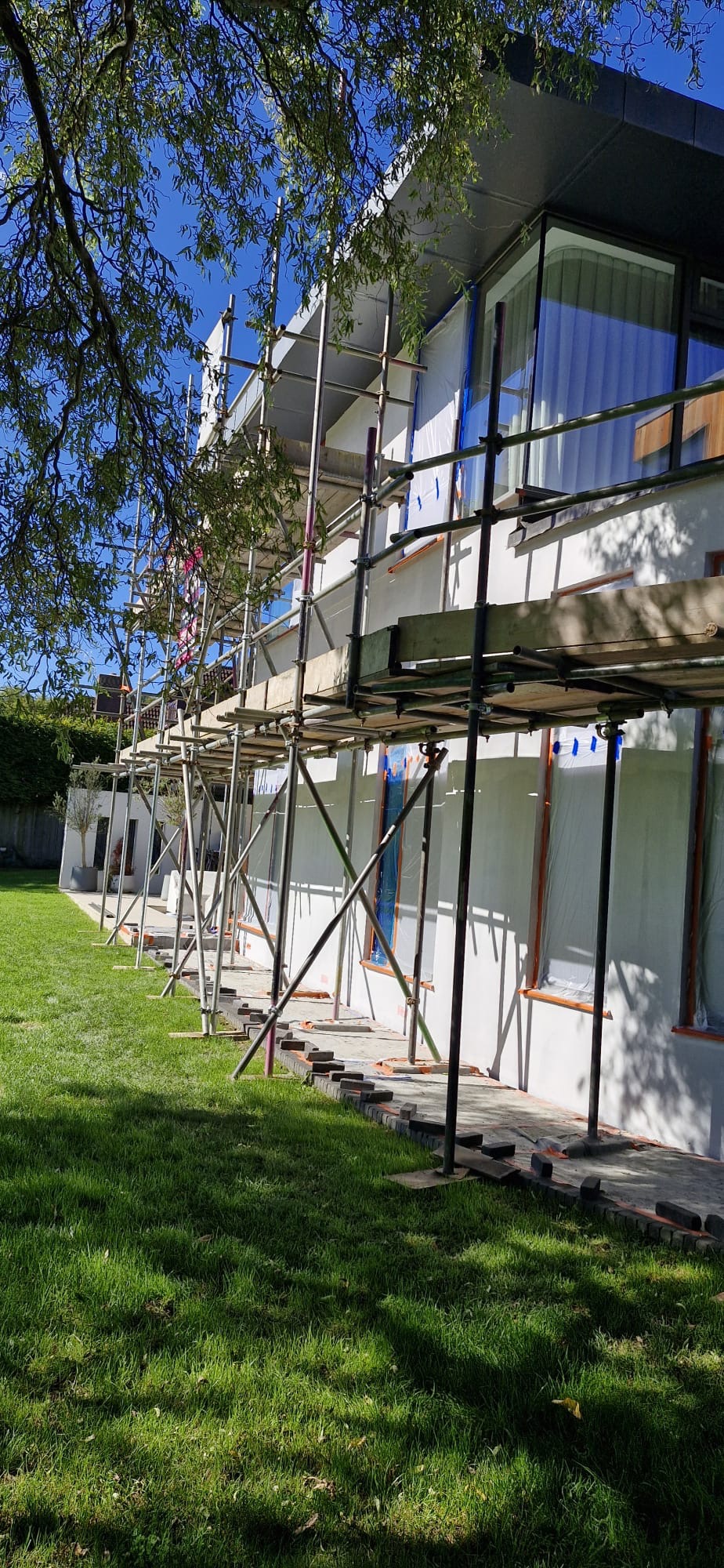 Dom jednorodzinny w trakcie prac elewacyjnych, z widocznymi rusztowaniami i zabezpieczeniami okien folią. Świeżo położony tynk na elewacji. Widok z poziomu trawnika.