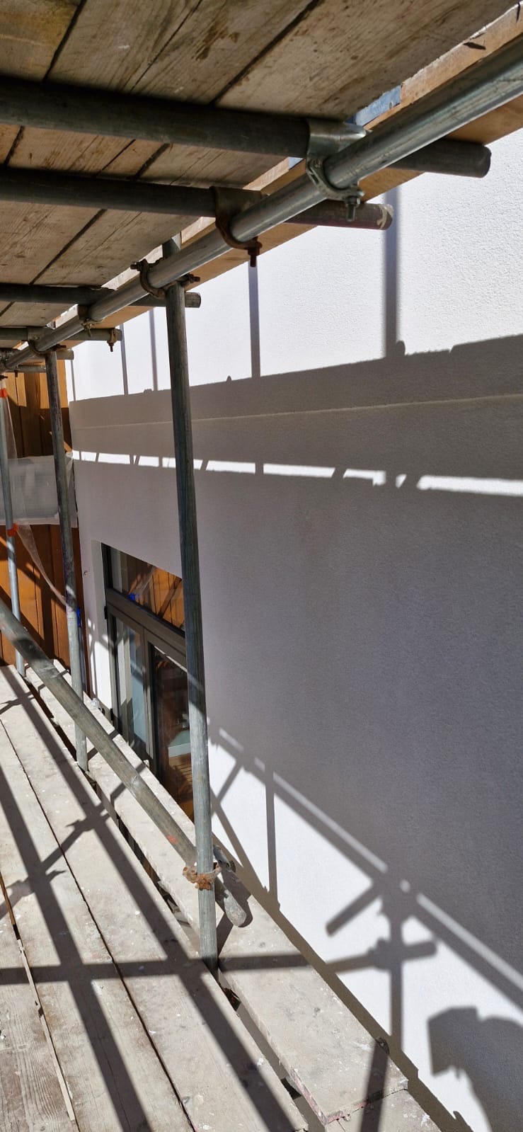 Biała elewacja budynku w trakcie prac, widoczna z poziomu rusztowania. Cień konstrukcji na ścianie, okno w tle. Ujęcie z dołu, podkreślające wysokość.