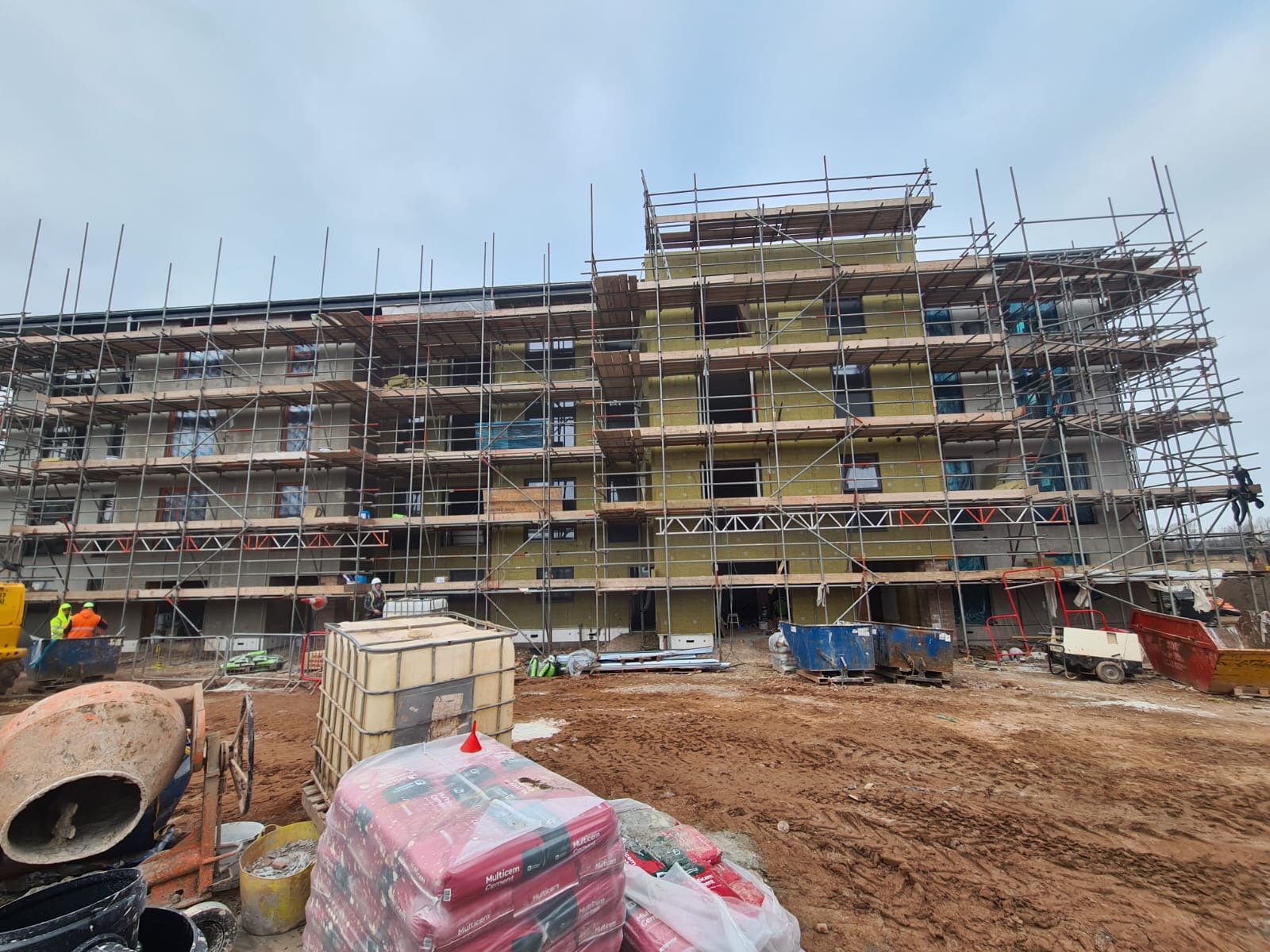 Budynek wielorodzinny w trakcie ocieplania, widoczne rusztowania, materiały budowlane i ekipa w kaskach. Etap prac: izolacja ścian zewnętrznych.