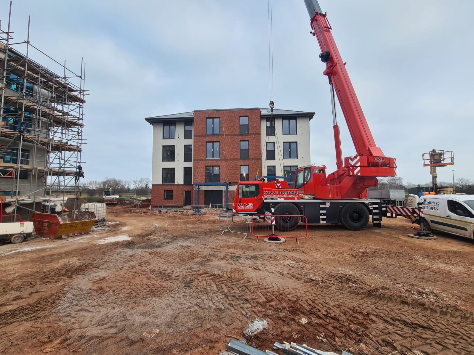 Budynek w trakcie wykończenia elewacji z użyciem dźwigu, widoczne rusztowania i teren budowy. Część fasady wykończona czerwoną cegłą, reszta w jasnym tynku.