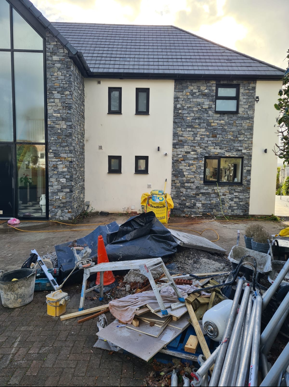 Dom z elewacją z kamienia, widoczne narzędzia i materiały budowlane. Fragment ukończonej elewacji z kamienia, okna w czarnych ramach. Częściowy widok domu.