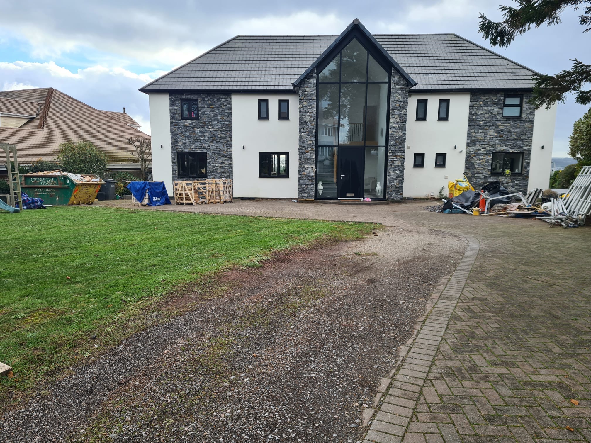 Dwukondygnacyjny dom z elewacją z kamienia i białego tynku, z dużym przeszkleniem nad wejściem. Na trawniku widoczne materiały budowlane i kontener.