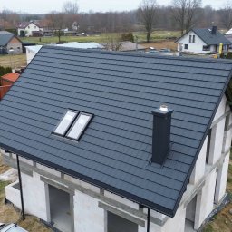 Dachy NP - Nowoczesny, grafitowy dach z dwoma oknami połaciowymi i kominem na surowym budynku w trakcie budowy. Widok z góry ukazuje precyzję krycia.