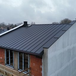 Dachy NP - Nowoczesny, czarny dach z blachy na budynku w trakcie budowy, widok z góry. Częściowo otynkowana ściana, okna bez parapetów, rusztowanie. Pochmurne niebo w tle.