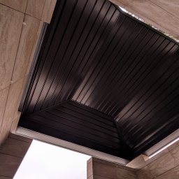 Dachy NP - Nowoczesne zadaszenie z ciemnej blachy trapezowej, widok z dołu, z elementami wykończenia z płytek imitujących kamień. Minimalistyczny design.