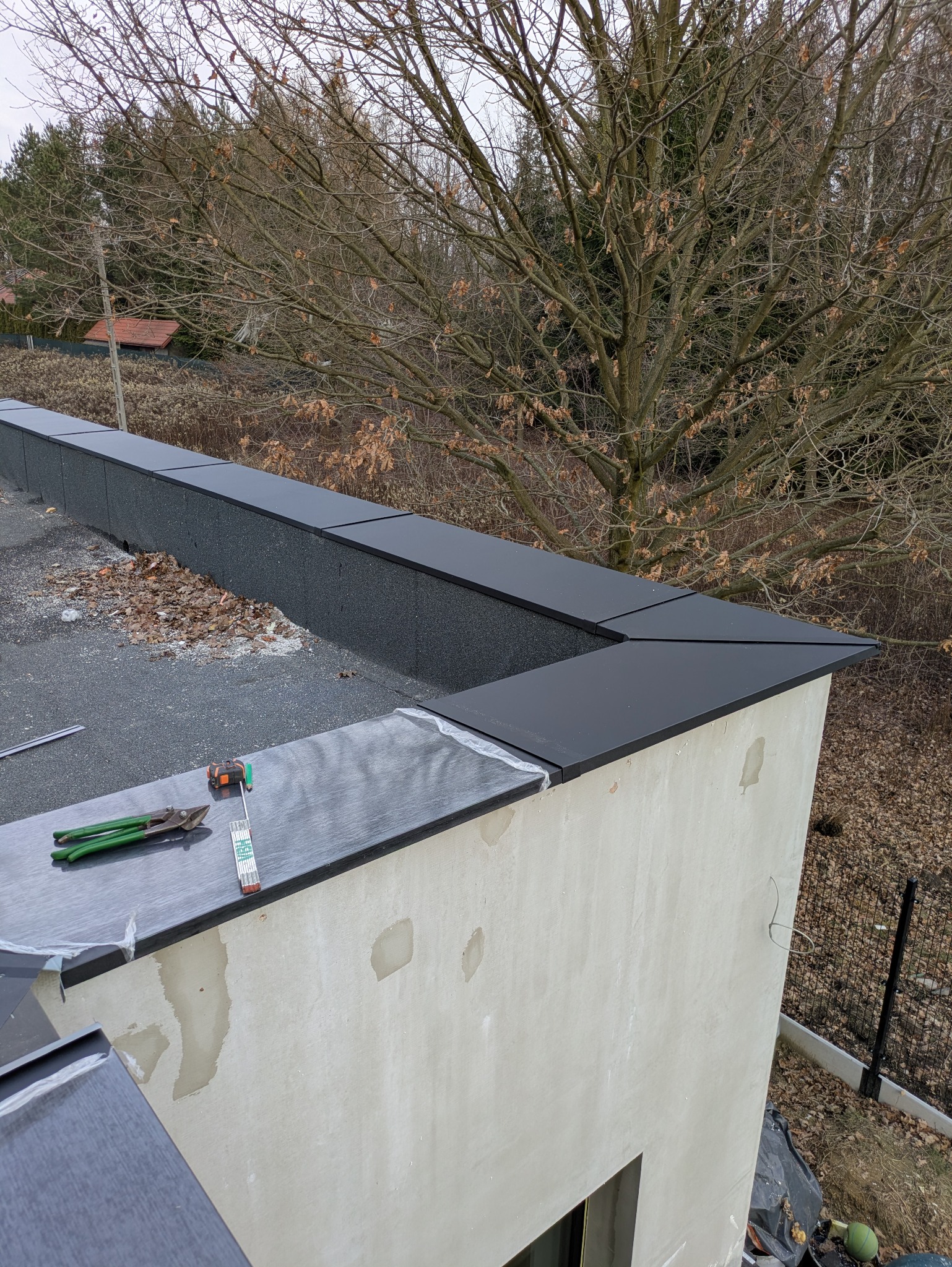 Wykończony dach płaski z czarnymi obróbkami blacharskimi na krawędziach. Widoczne narzędzia dekarskie i fragment elewacji budynku. Tło stanowią drzewa i zabudowa.