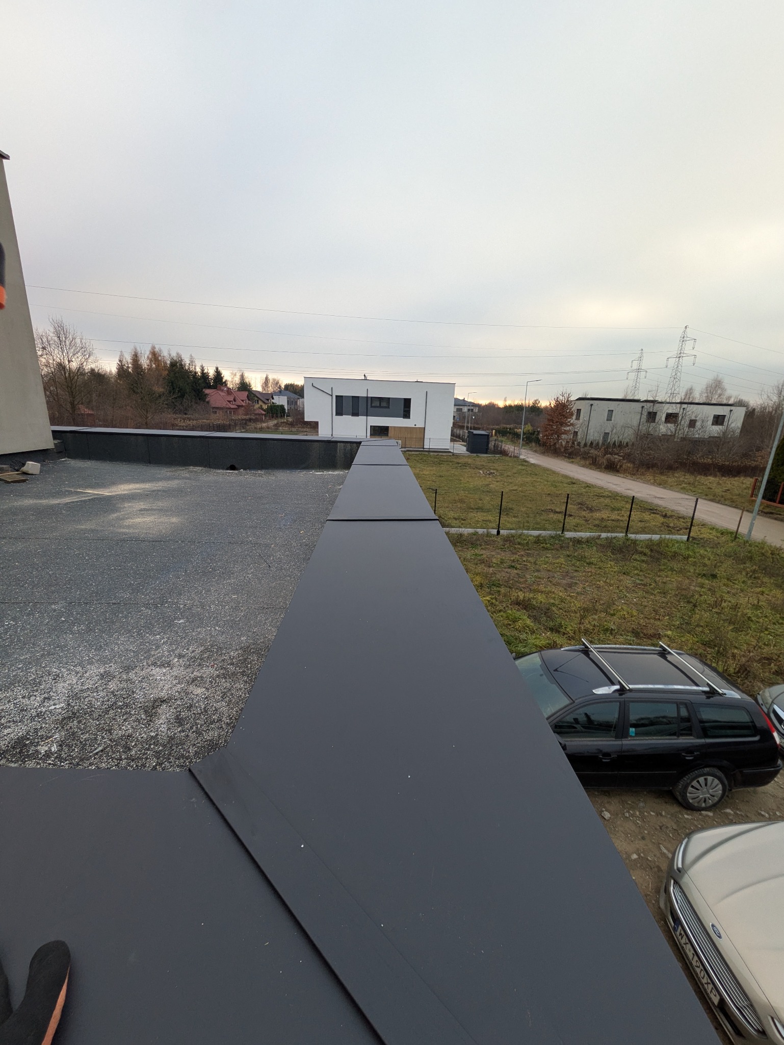 Nowoczesny, płaski dach pokryty czarną blachą na tle zabudowy jednorodzinnej i pochmurnego nieba. Widok z góry, ukazujący precyzję wykonania obróbek blacharskich.