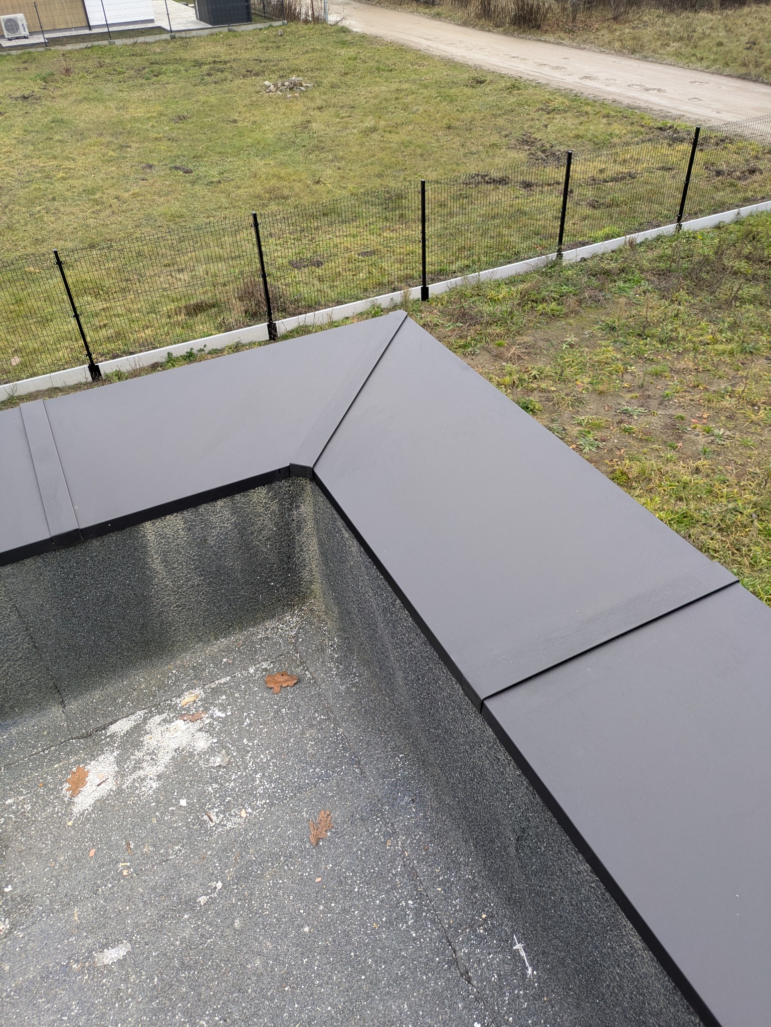 Szczegółowe ujęcie płaskiego dachu pokrytego czarną papą, z widocznymi obróbkami blacharskimi i fragmentem zielonego trawnika w tle. Widok z góry.