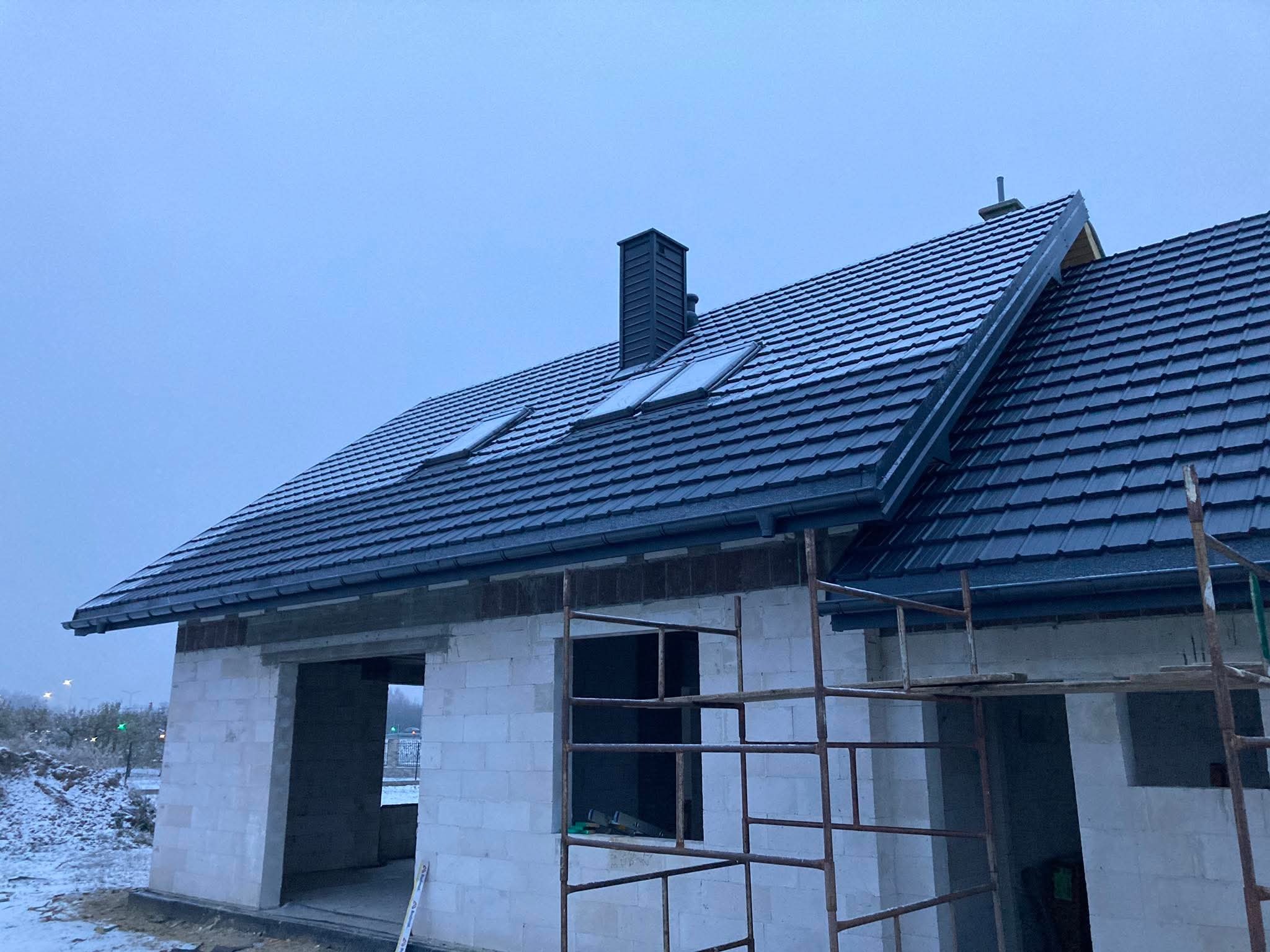 Dom w trakcie budowy z ciemnym dachem pokrytym dachówką, widoczne okna dachowe i komin. Częściowo ośnieżony teren i rusztowanie przy ścianie.