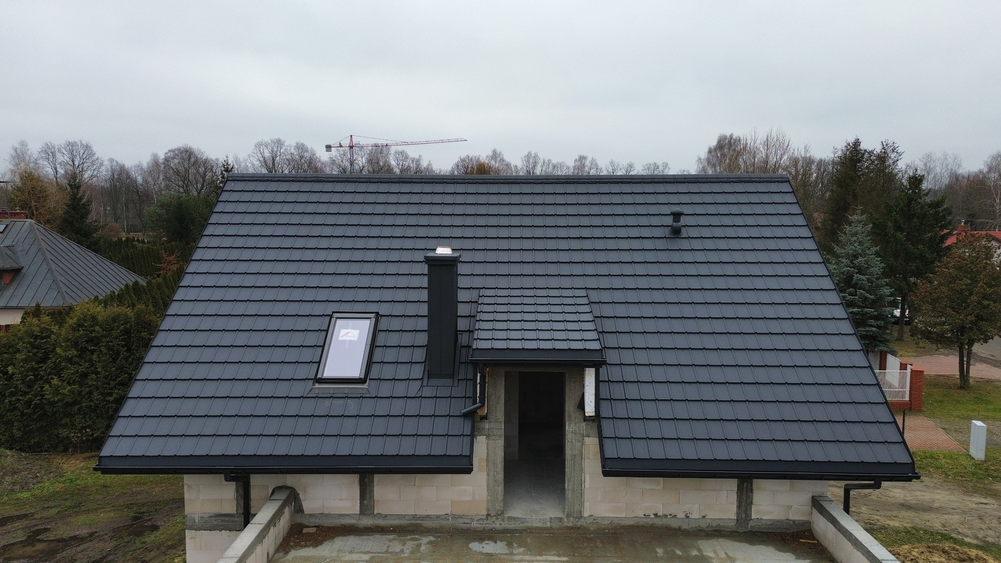 Grafitowy dach z dachówki modułowej, okno dachowe, komin i rynny w kolorze czarnym. Budynek w trakcie budowy, widoczny żuraw budowlany w tle.