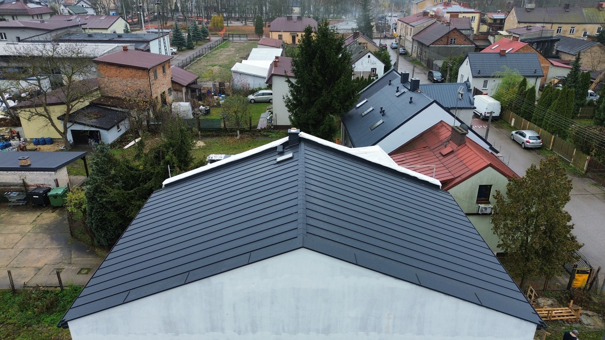 Nowoczesny, grafitowy dach z blachy na białym budynku, widok z góry. W tle zabudowa jednorodzinna i drzewa. Charakterystyczne kominy i wykończenia.
