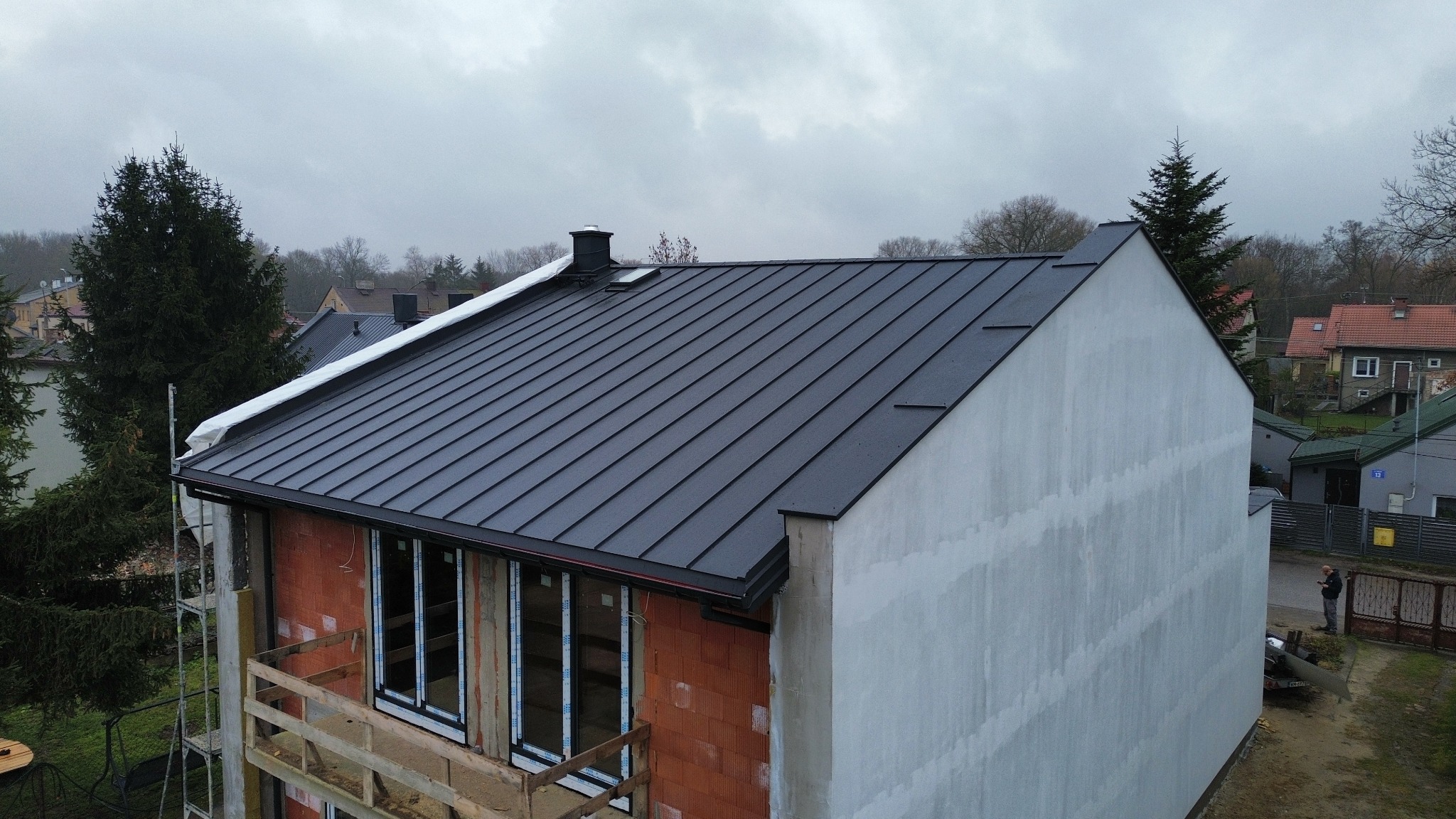 Nowoczesny, czarny dach z blachy na budynku w trakcie budowy, widok z góry. Częściowo otynkowana ściana, okna bez parapetów, rusztowanie. Pochmurne niebo w tle.