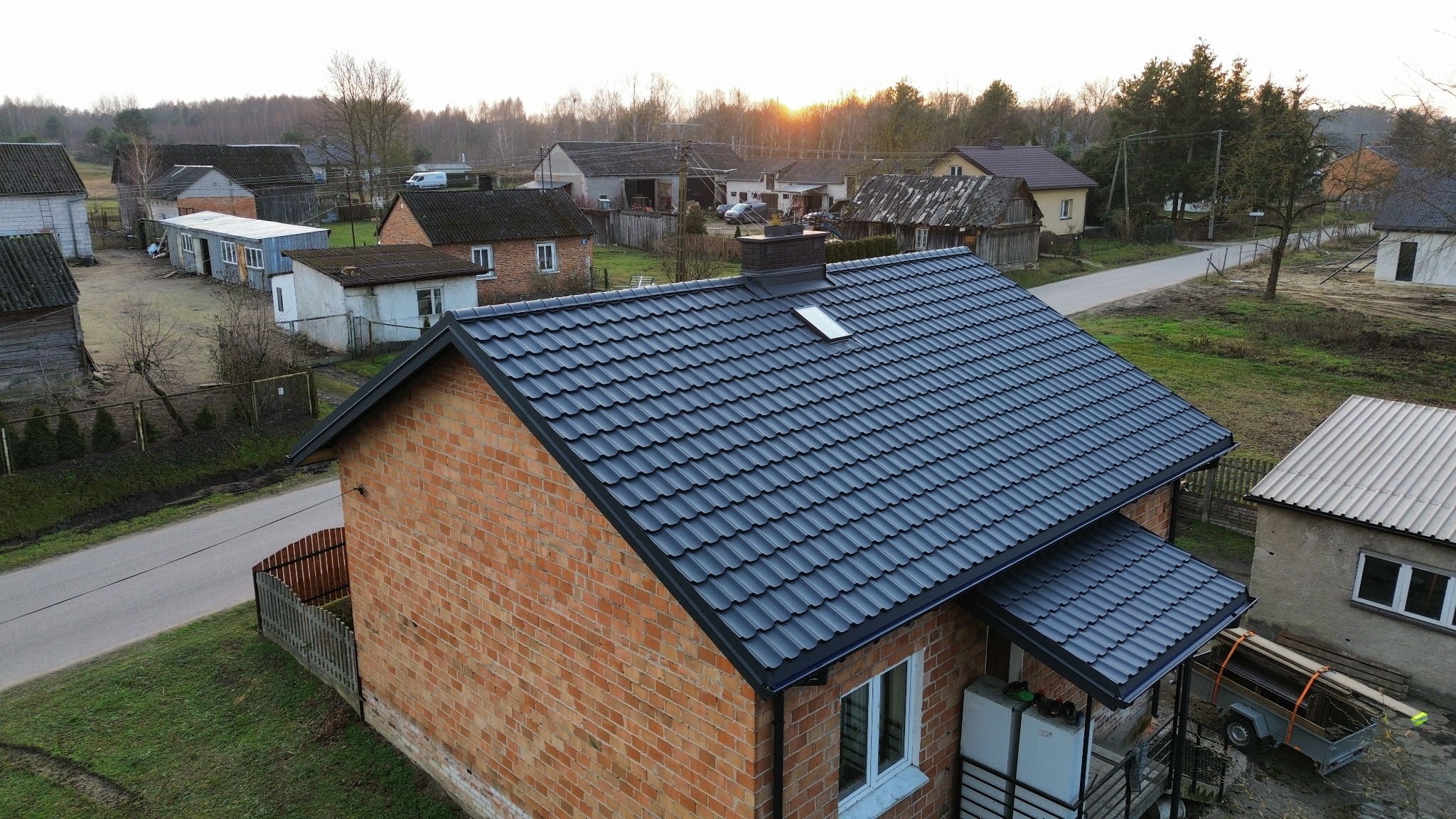 Ciemnoszary dach z blachodachówki z oknem dachowym na ceglanym domu jednorodzinnym, widok z góry na tle wiejskiej zabudowy o zmierzchu.