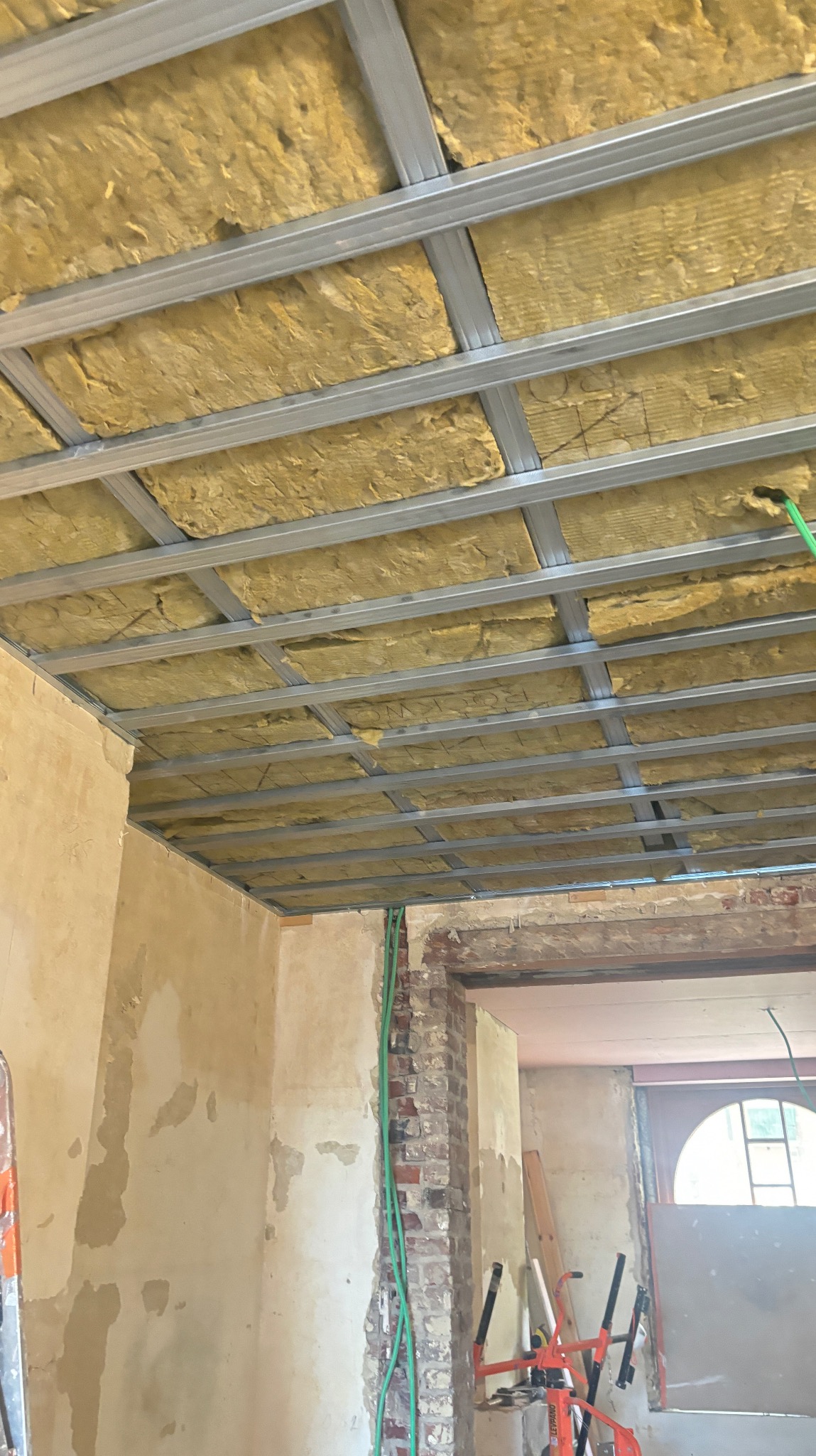 Montaż izolacji z wełny mineralnej w metalowym stelażu sufitu podwieszanego, widok z dołu. Fragment ściany z instalacją elektryczną i ceglanym obramowaniem drzwi.