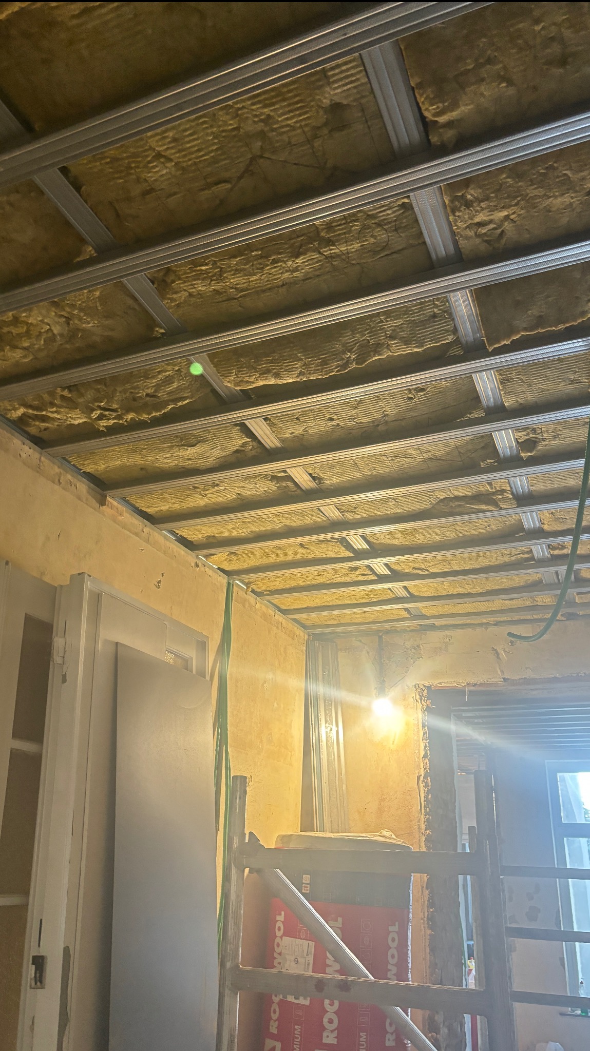 Montaż izolacji z wełny mineralnej na suficie podwieszanym z metalowym rusztem. Widoczne pakiety wełny i drabina. Prace remontowe w pomieszczeniu.