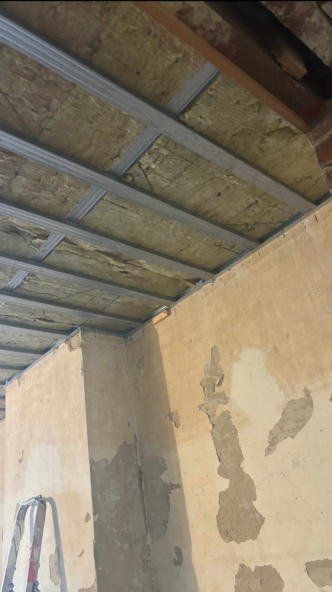 Ocieplenie poddasza wełną mineralną z widoczną konstrukcją metalową. Ściany w stanie surowym, widoczna drabina. Ujęcie z dołu, podkreślające etap prac.