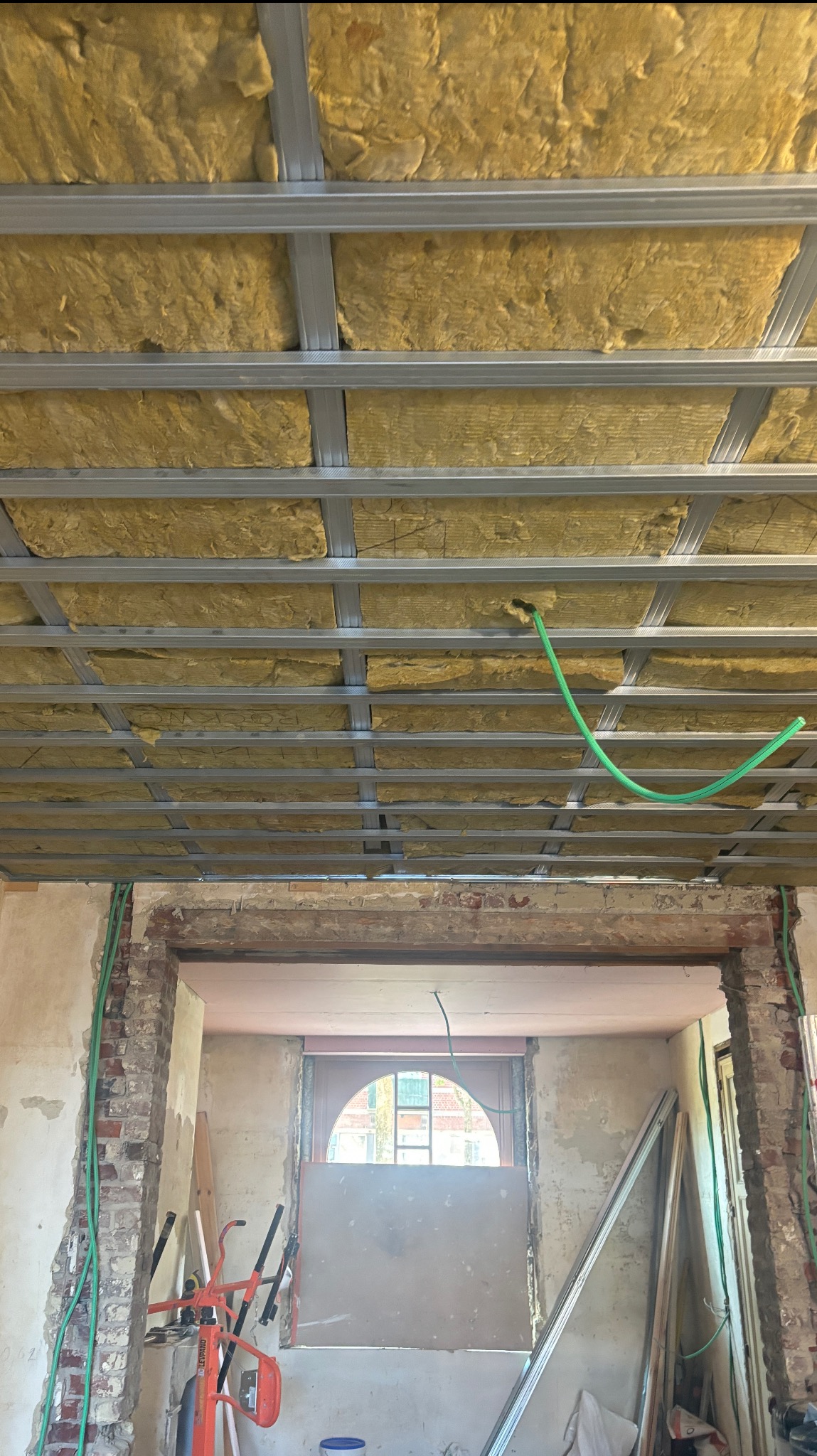 Sufit podwieszany z izolacją z wełny mineralnej, widoczne profile metalowe i instalacja. Fragment ściany z cegły i okno w tle. Prace remontowe w trakcie.