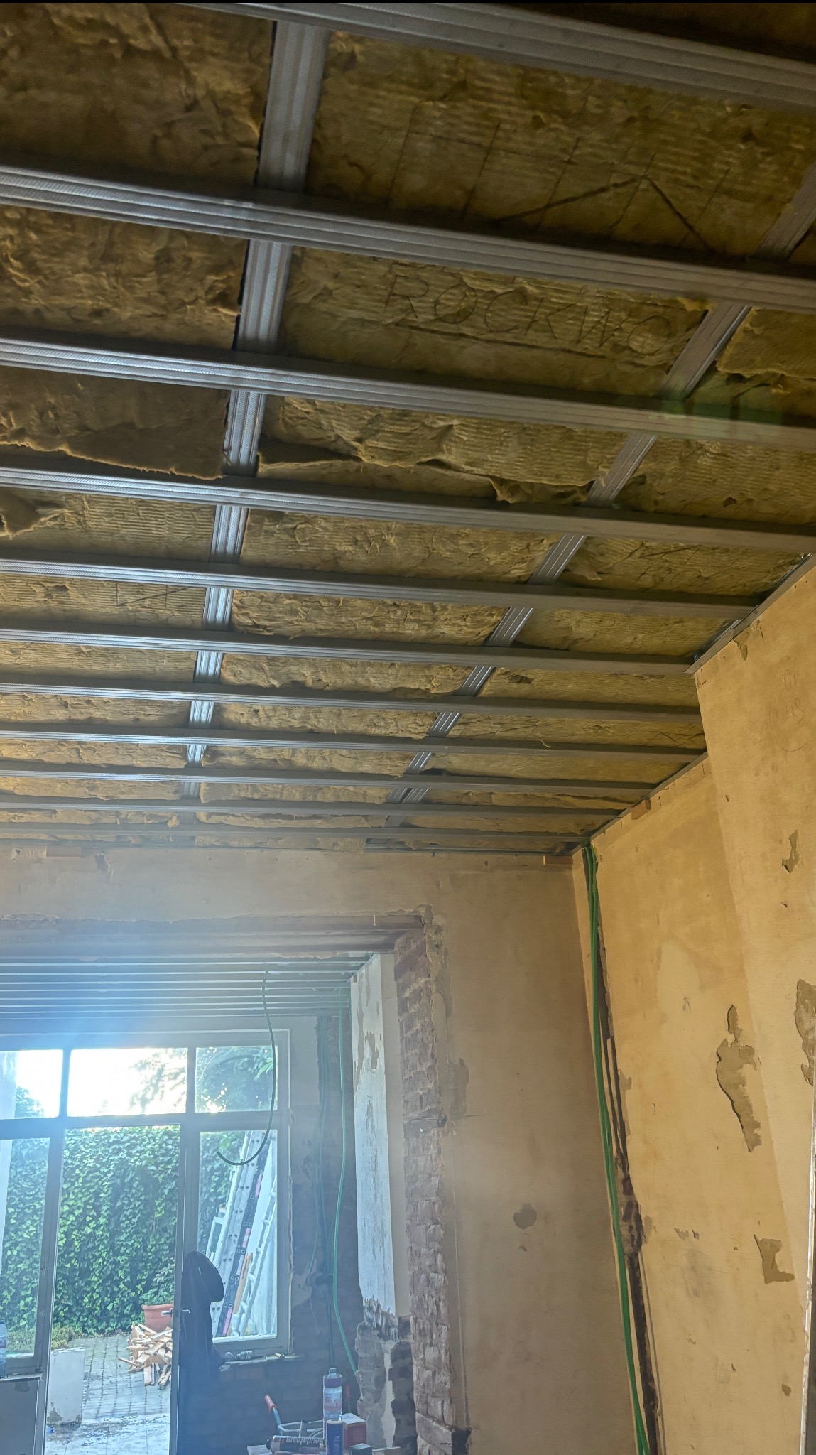 Montaż izolacji poddasza z wełny mineralnej Rockwool, widoczne metalowe profile konstrukcyjne. Ściany w stanie surowym, okno z widokiem na zieleń.