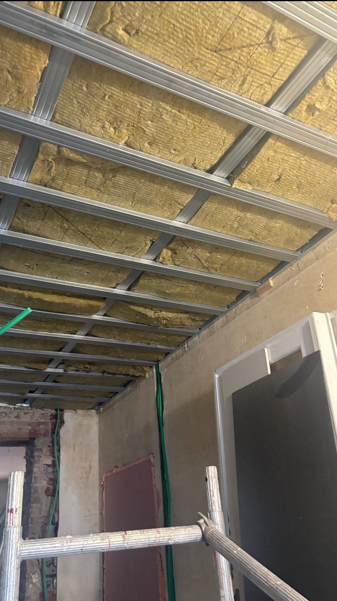 Montaż izolacji poddasza z wełny mineralnej w metalowym stelażu. Widoczne przewody elektryczne i fragment rusztowania. Prace remontowe w trakcie realizacji.