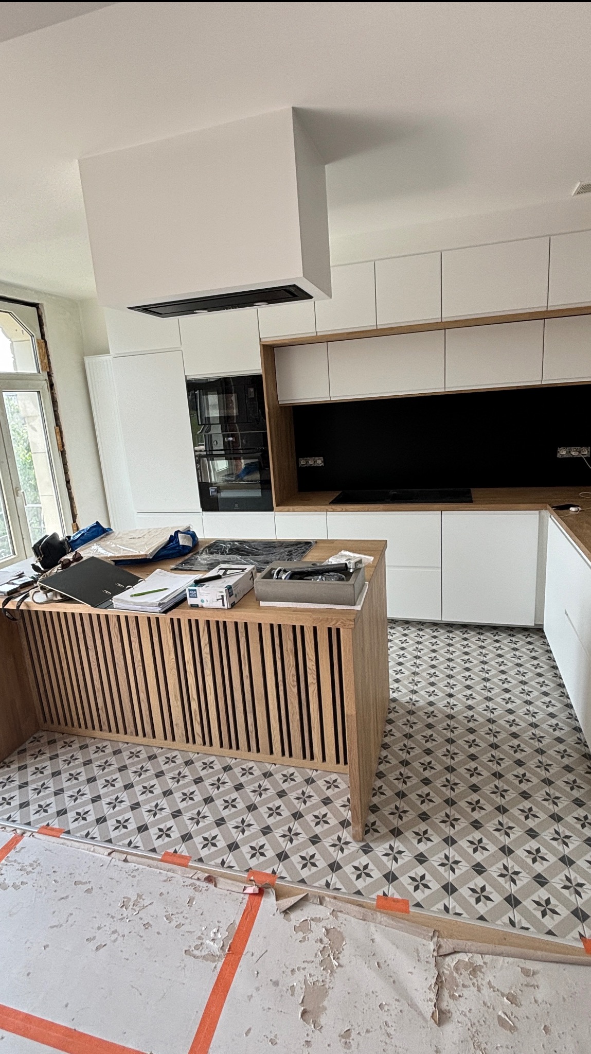 Nowoczesna kuchnia w trakcie wykończenia: białe szafki, drewniana wyspa z pionowymi listwami i wzorzysta podłoga. Widoczne zabezpieczenia podłogi.