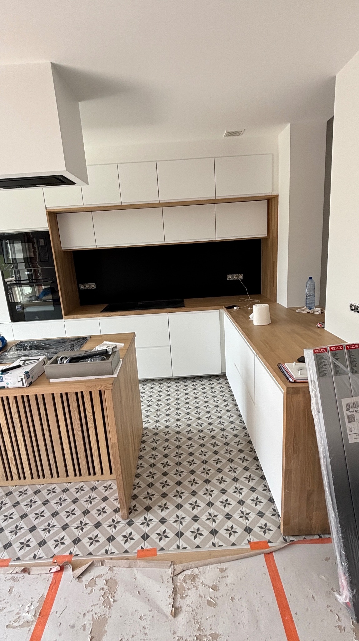 Nowoczesna kuchnia z białą zabudową i drewnianymi akcentami. Na podłodze wzorzyste płytki. Widoczne elementy wykończeniowe i zabezpieczenia podłogi.