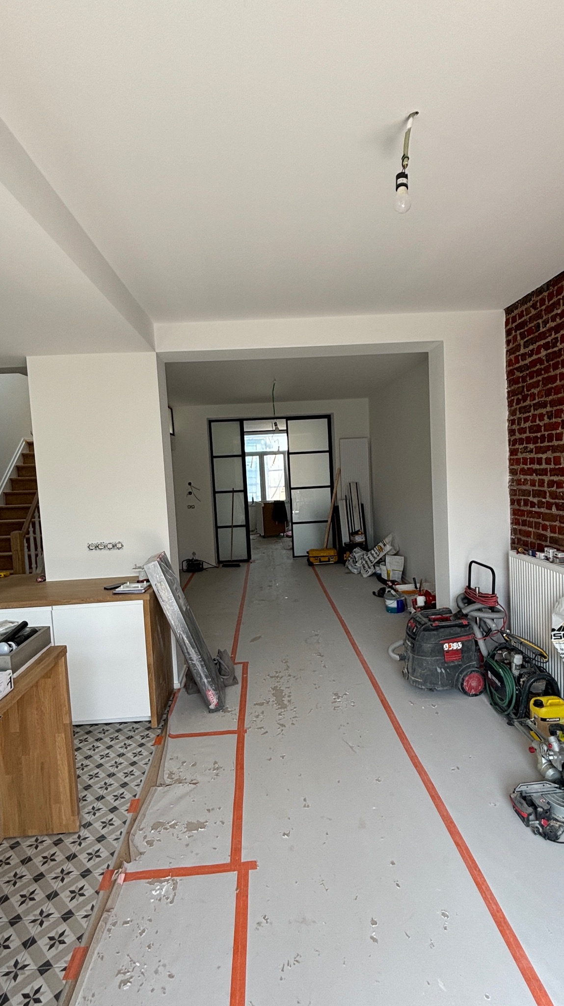 Wnętrze w trakcie remontu: zabezpieczona podłoga, narzędzia budowlane, widoczna ceglana ściana i przeszklone drzwi przesuwne. Etap prac wykończeniowych.