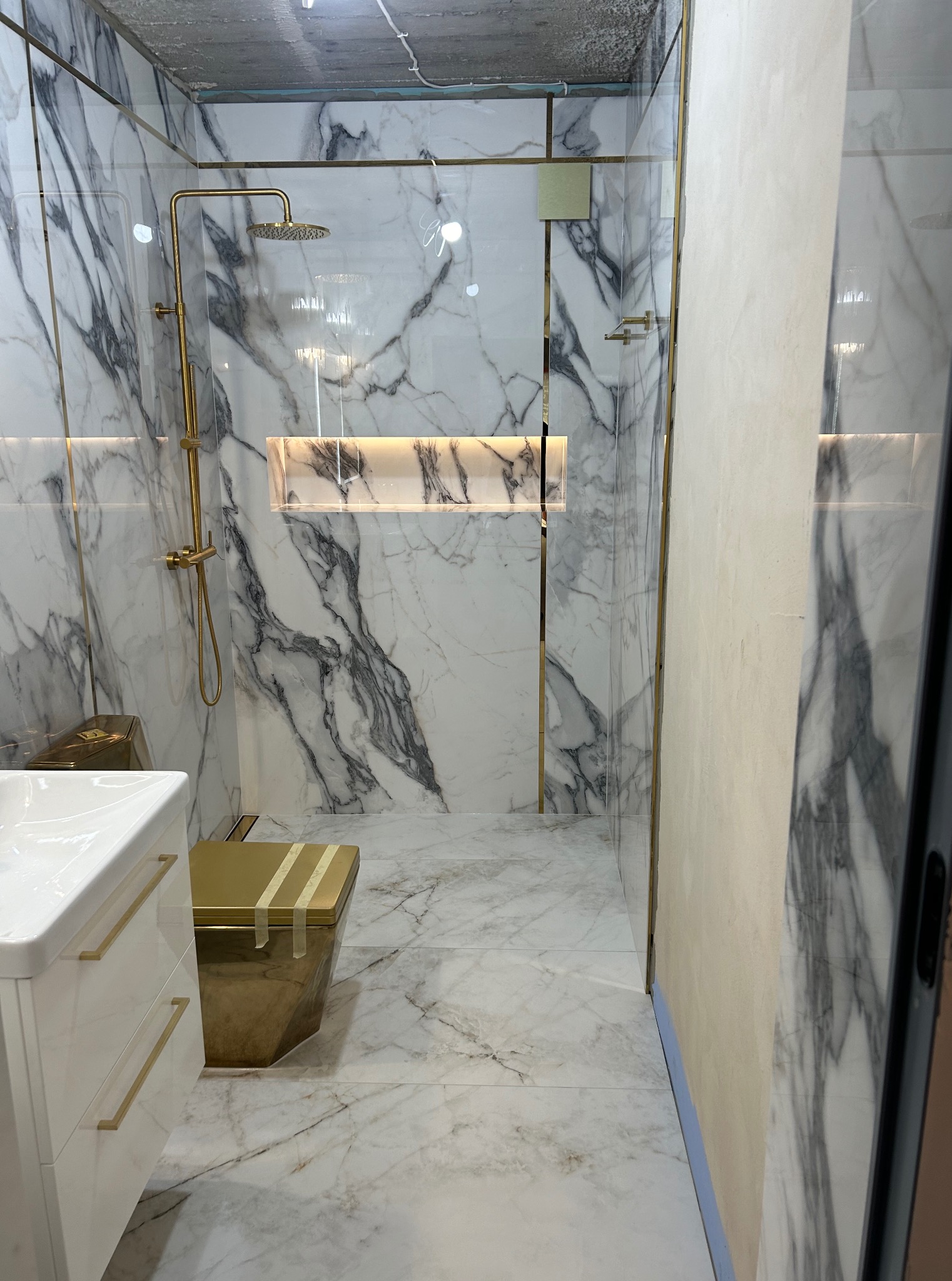 Wykończona łazienka z marmurowymi płytkami, złotą armaturą i kabiną prysznicową typu walk-in. Widoczna umywalka z szafką i toaleta zabezpieczona folią.