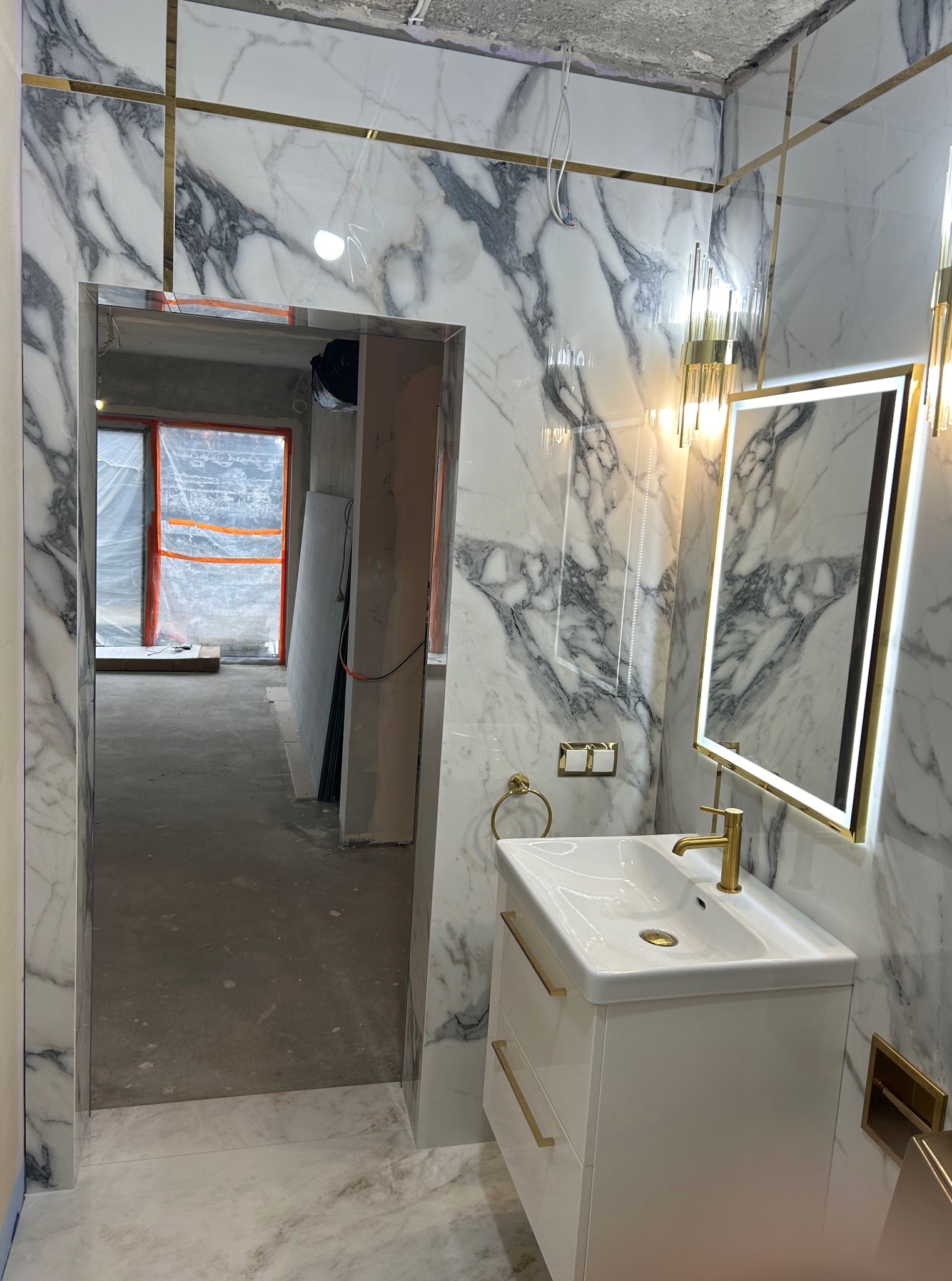 Wykończona łazienka z marmurowymi płytkami, złotymi akcentami i nowoczesną umywalką. Widok na surowy stan drugiego pomieszczenia przez otwarte drzwi.