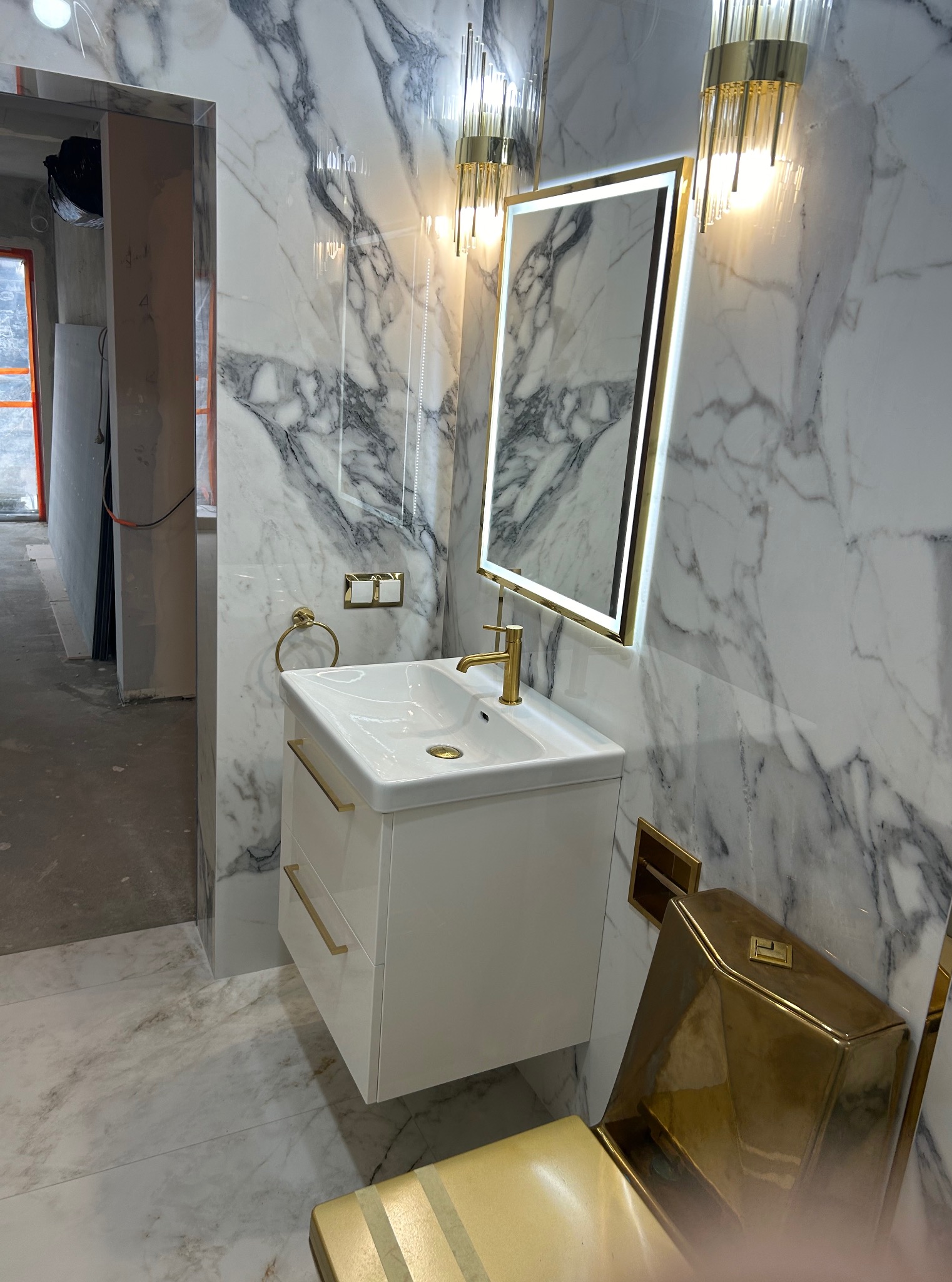 Wykończona łazienka z marmurowymi płytkami, białą szafką umywalkową i złotymi akcentami, w tym armaturą i oświetleniem. Widoczny fragment toalety w złotym kolorze.