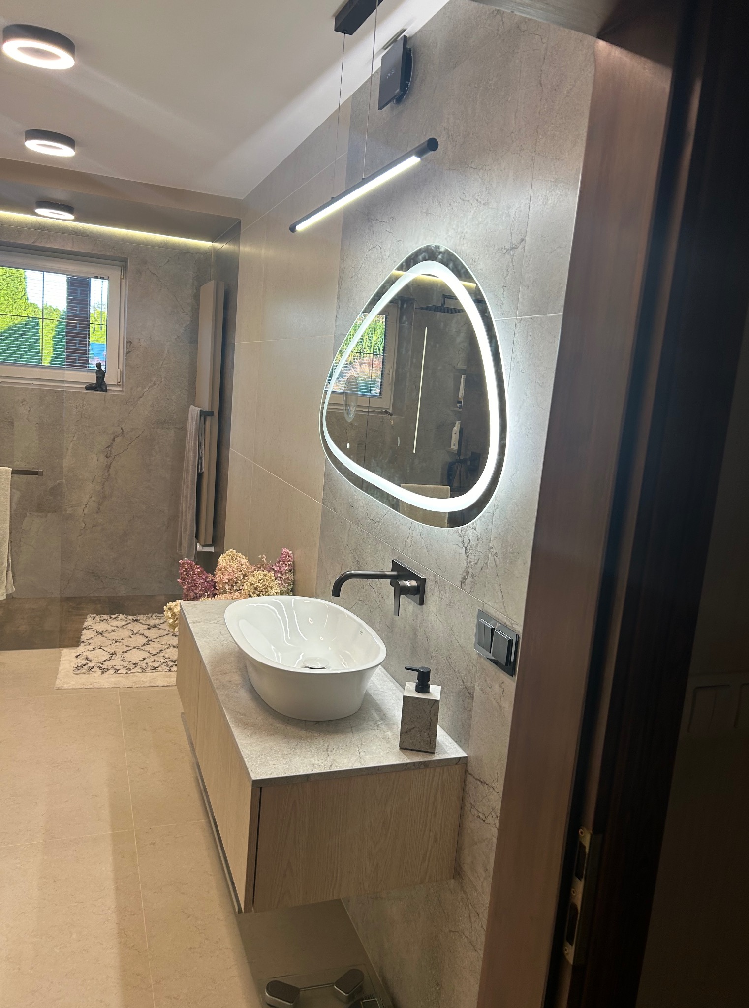 Nowoczesna łazienka z owalną umywalką nablatową, podświetlanym lustrem i płytkami imitującymi marmur. Minimalistyczny design, ciepłe oświetlenie, widok na zieleń za oknem.