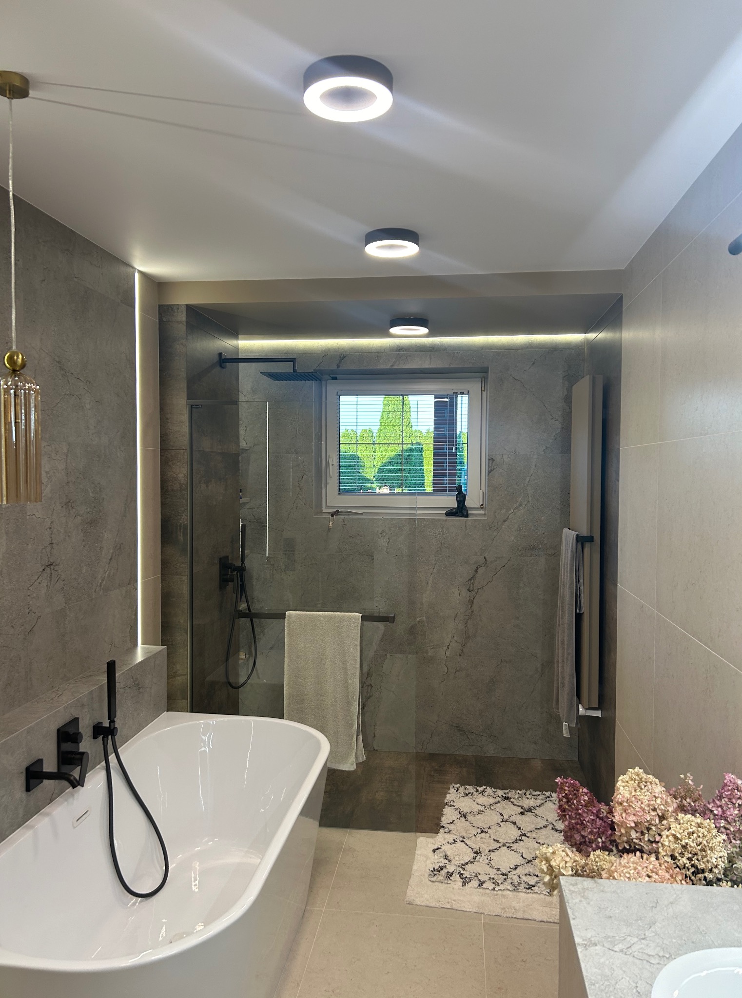 Nowoczesna łazienka z szarymi płytkami imitującymi kamień, białą wanną i czarną armaturą. Uwagę przykuwa dekoracyjny dywanik i bukiet kwiatów.