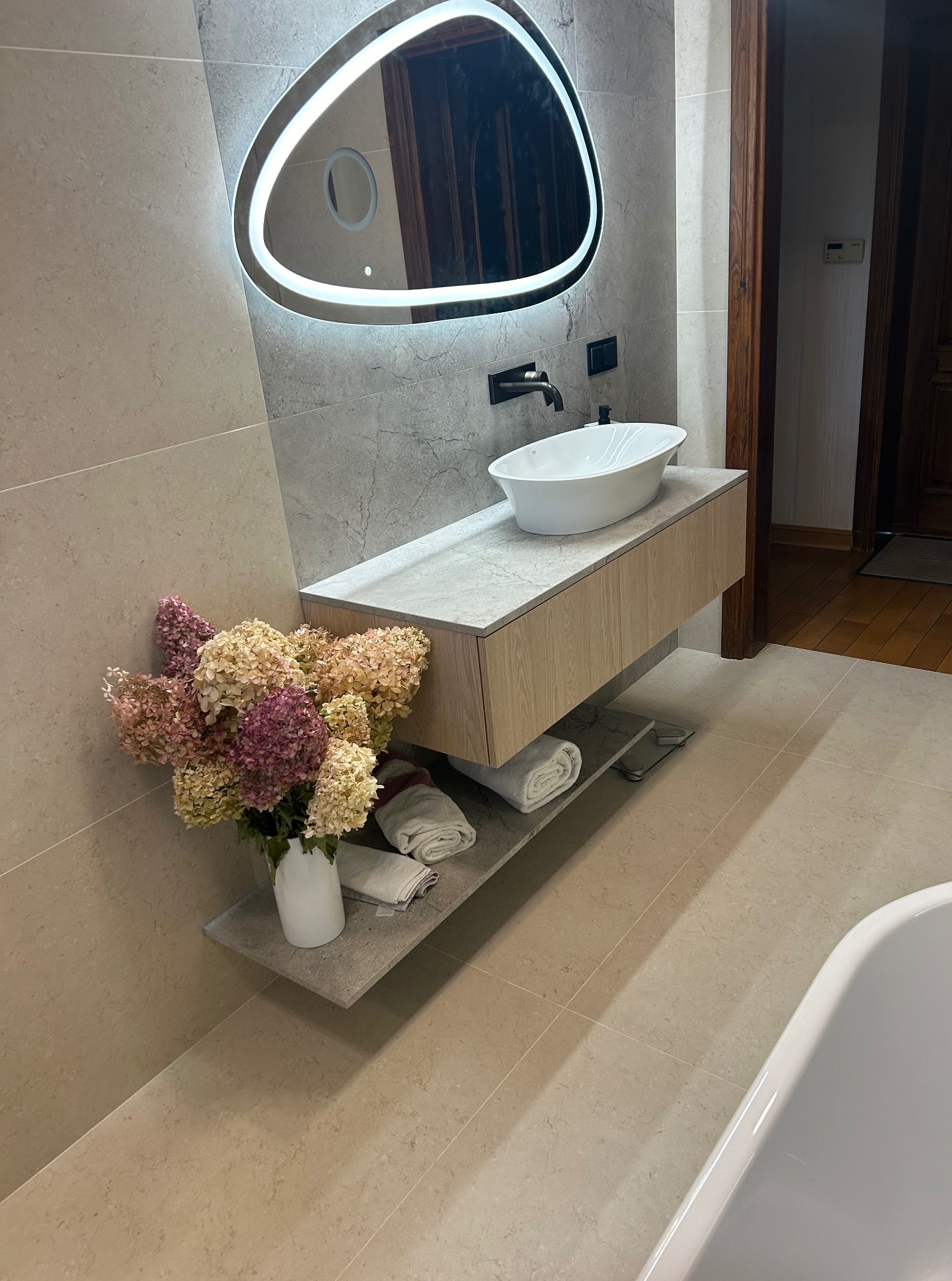 Elegancka łazienka w Gdyni z beżowymi płytkami, owalnym lustrem z podświetleniem LED i umywalką nablatową. Minimalistyczny design z kwiatami.