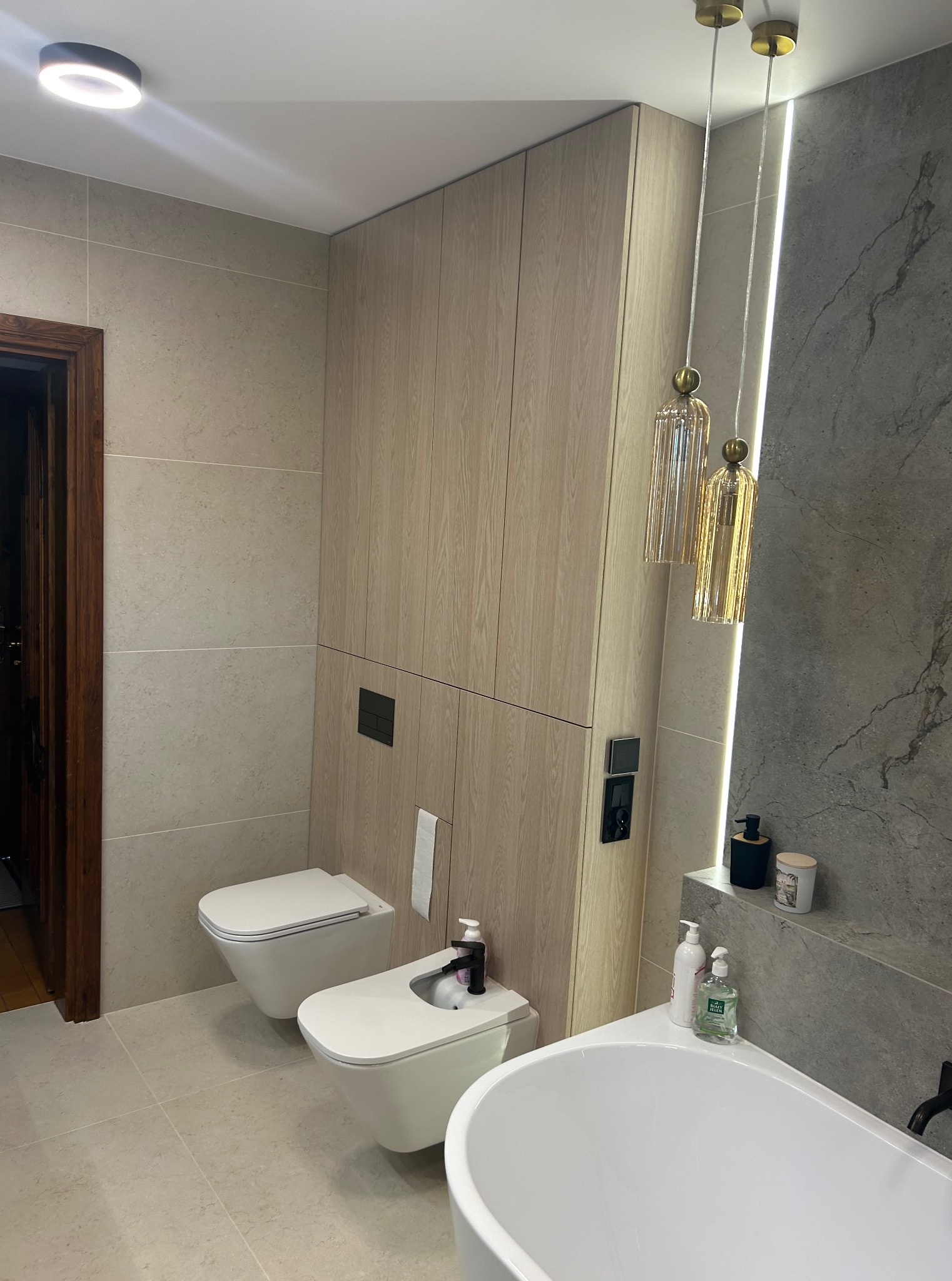 Nowoczesna łazienka z beżowymi płytkami, wiszącą toaletą i bidetem. Uwagę przykuwają złote lampy wiszące i drewniane szafki w zabudowie. Minimalistyczny design.