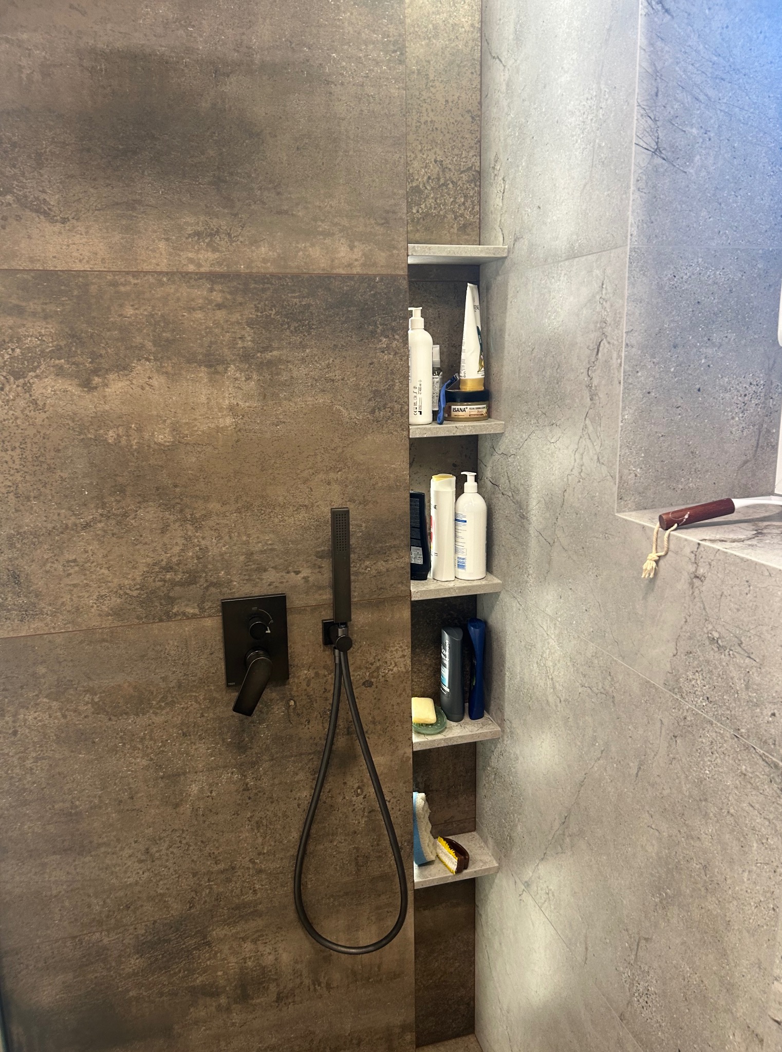 Nowoczesna kabina prysznicowa z ciemnobrązowymi i szarymi płytkami imitującymi kamień. Wnęka z półkami na kosmetyki i czarna armatura.