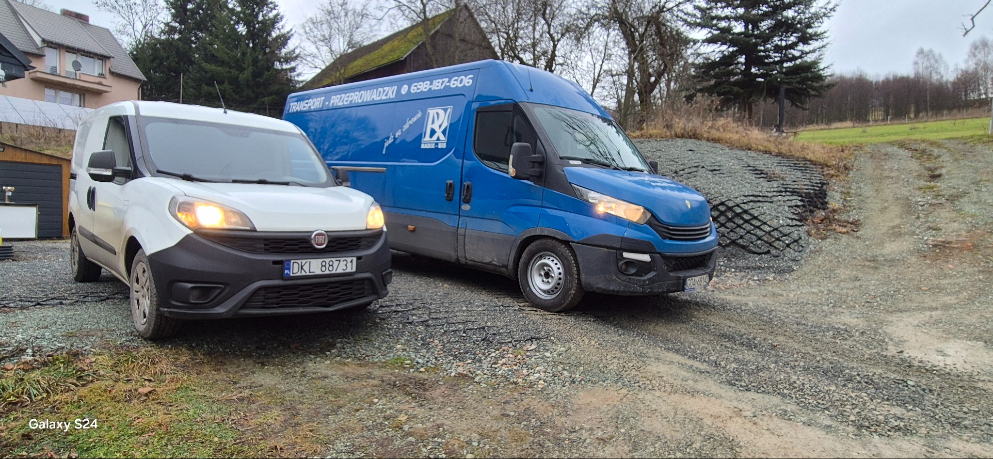 Dwa busy transportowe: biały Fiat Doblo i niebieski Iveco, stoją na żwirowej drodze. Na niebieskim busie widnieje napis 'TRANSPORT - PRZEPROWADZKI' i numer telefonu.