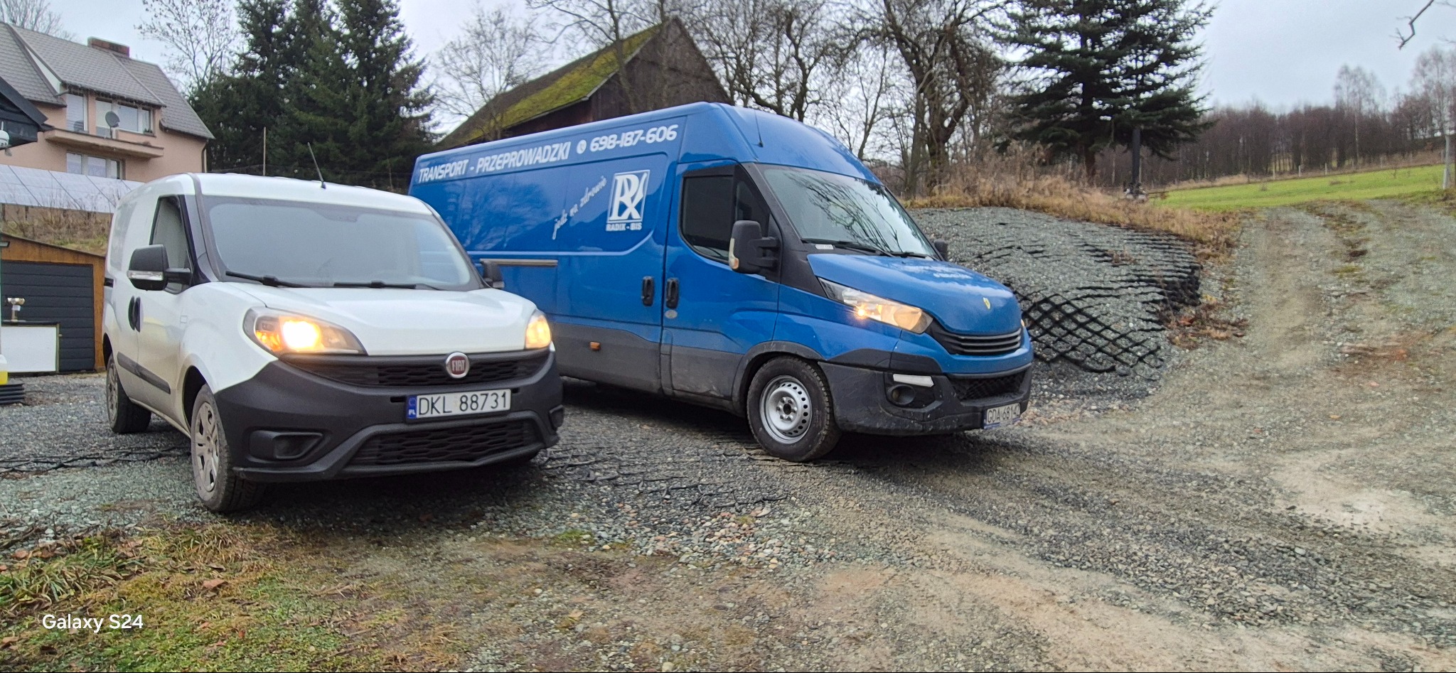 Dwa busy: biały Fiat Doblo z przodu i niebieski Iveco z napisem 'Transport - Przeprowadzki' w tle, stoją na żwirowej drodze w pochmurny dzień.