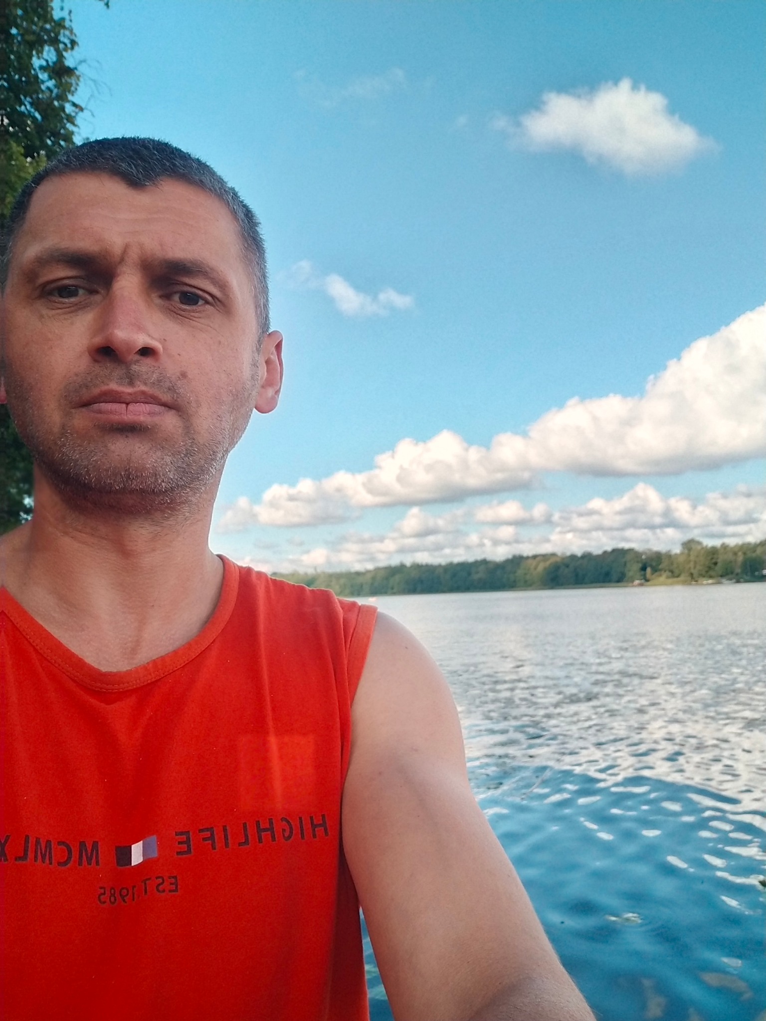 Mężczyzna w pomarańczowej koszulce na tle jeziora i błękitnego nieba z chmurami. Widoczny fragment drzewa po lewej stronie kadru. Ujęcie z bliska, skupione na twarzy.