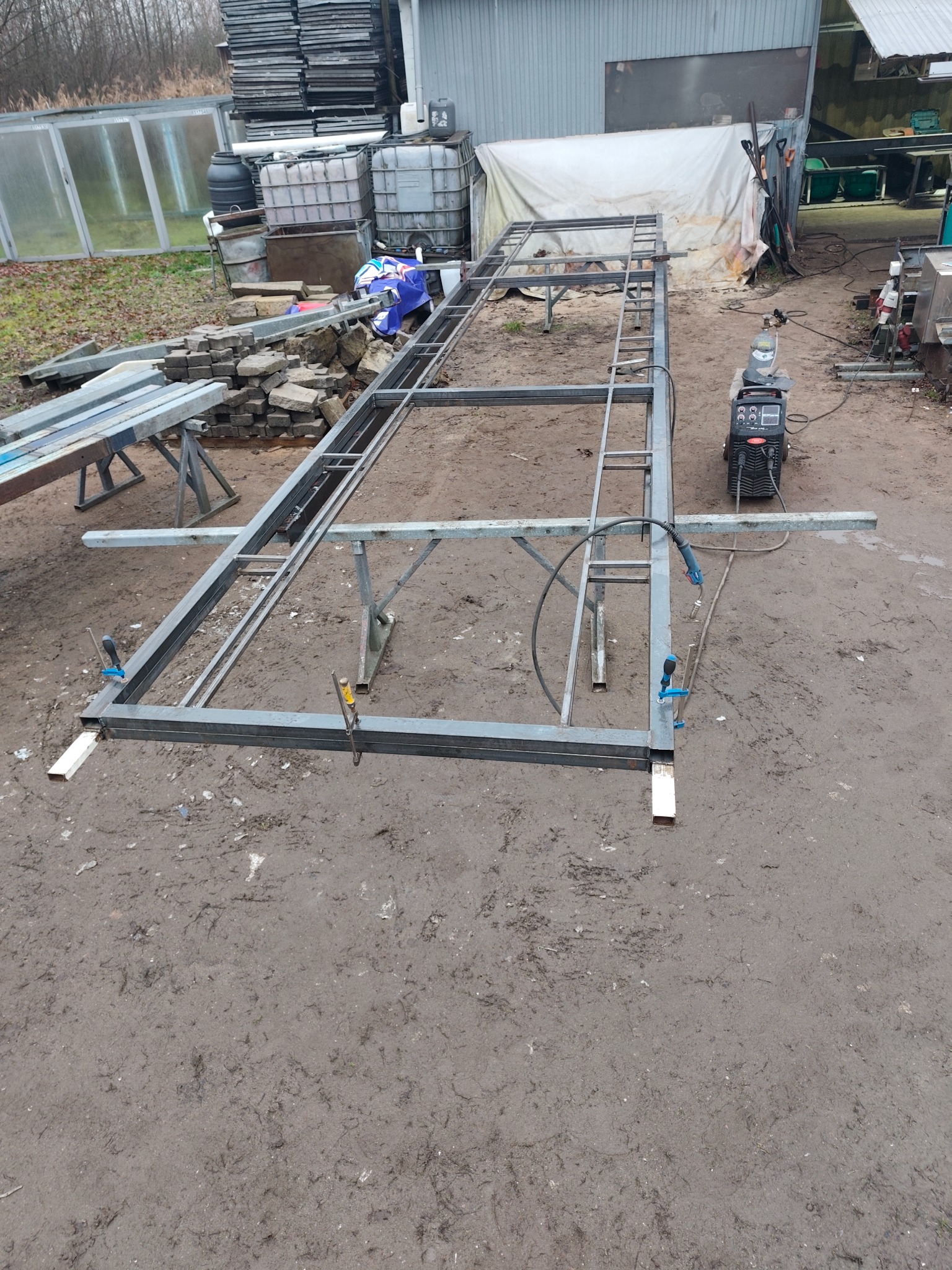 Konstrukcja metalowa w trakcie spawania na zewnątrz, widoczny sprzęt spawalniczy i elementy mocujące. Surowy teren budowy w tle.