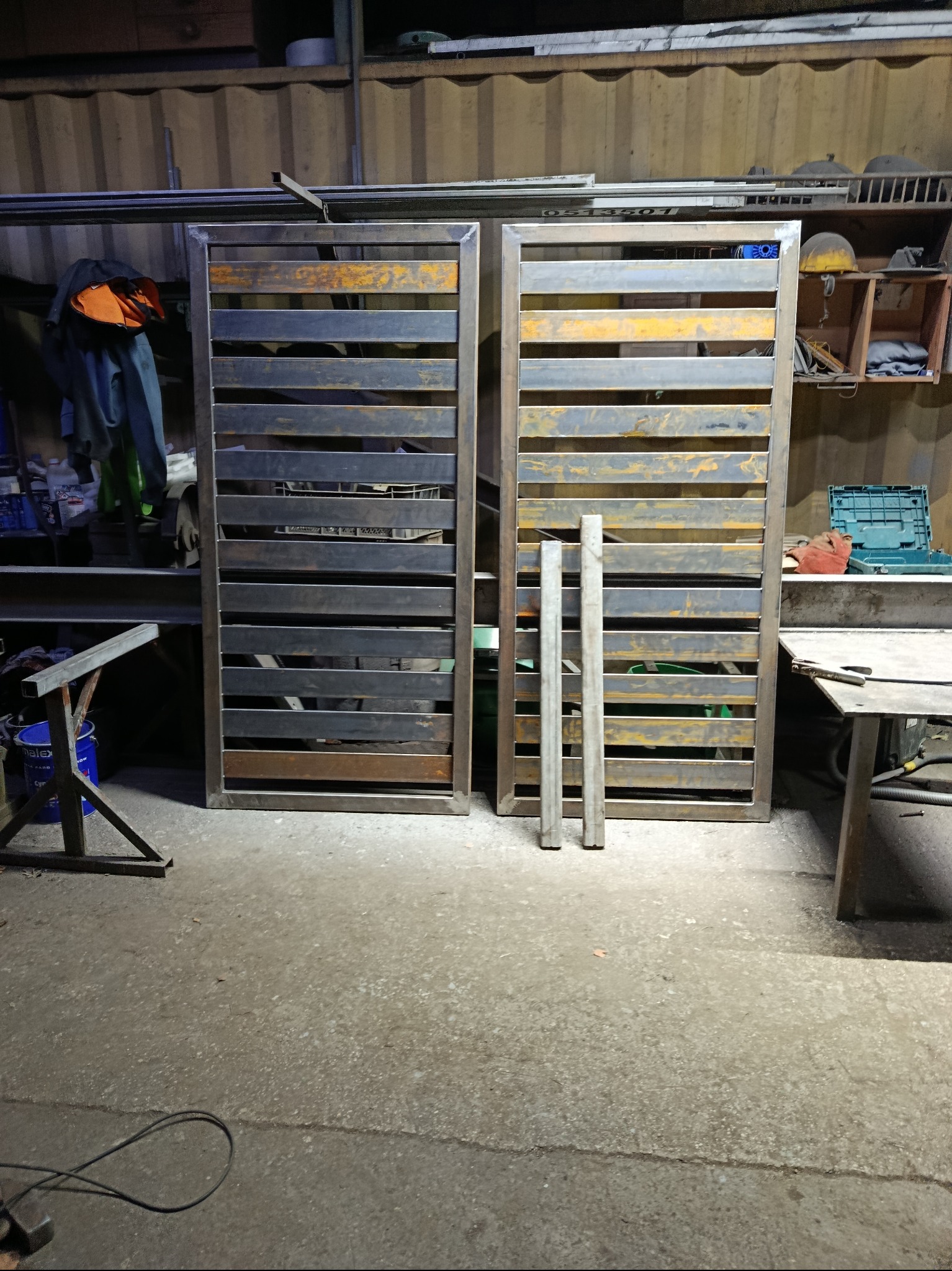 Dwie metalowe, surowe bramy z poziomymi szczeblami, w warsztacie. Widoczne ślady spawania i obróbki. Dwa metalowe słupki obok jednej z bram.