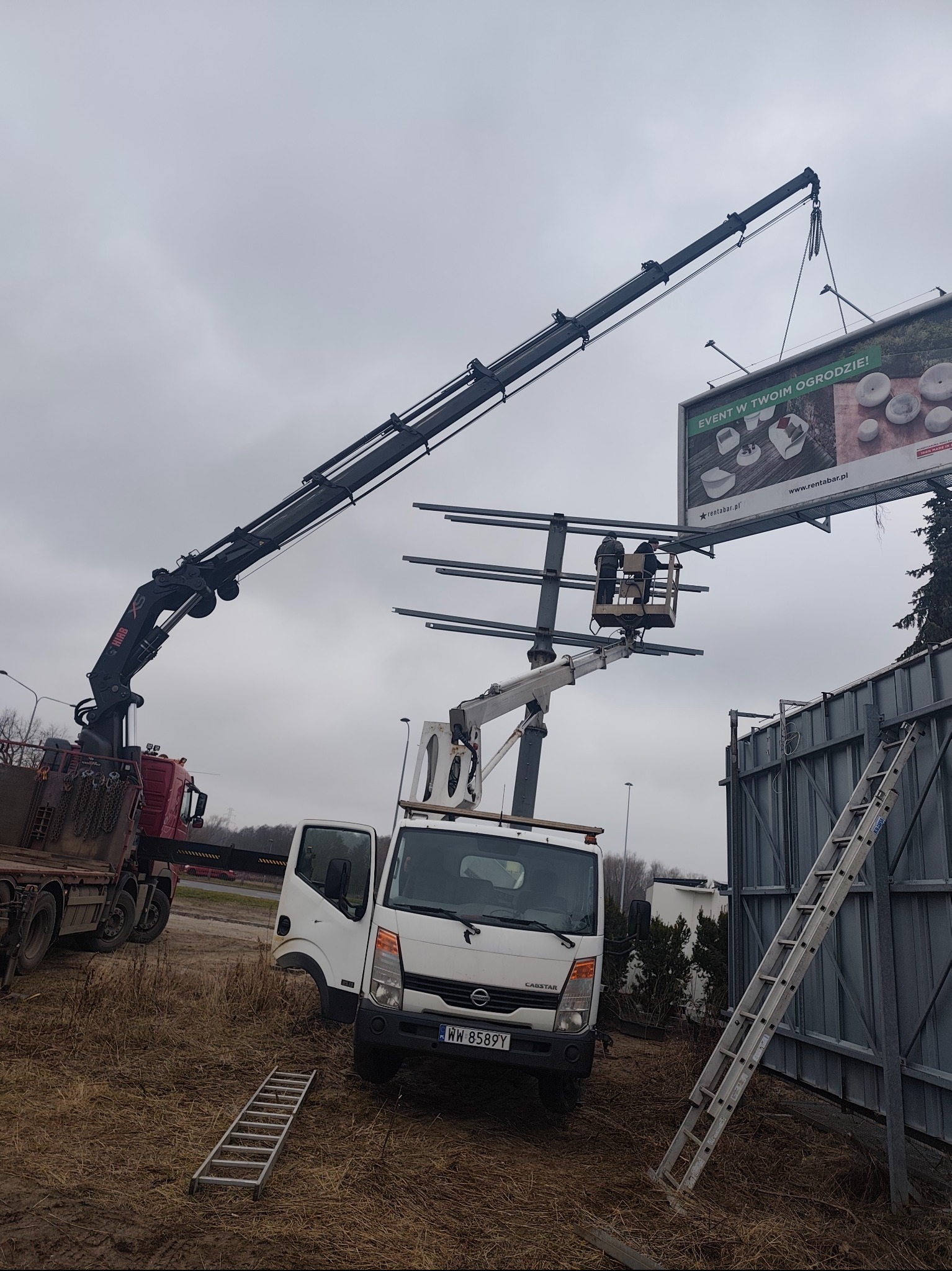 Montaż billboardu reklamowego z użyciem dźwigu i podnośnika koszowego. Widoczni pracownicy w trakcie pracy na wysokości. Konstrukcja metalowa i drabina oparta o nią.