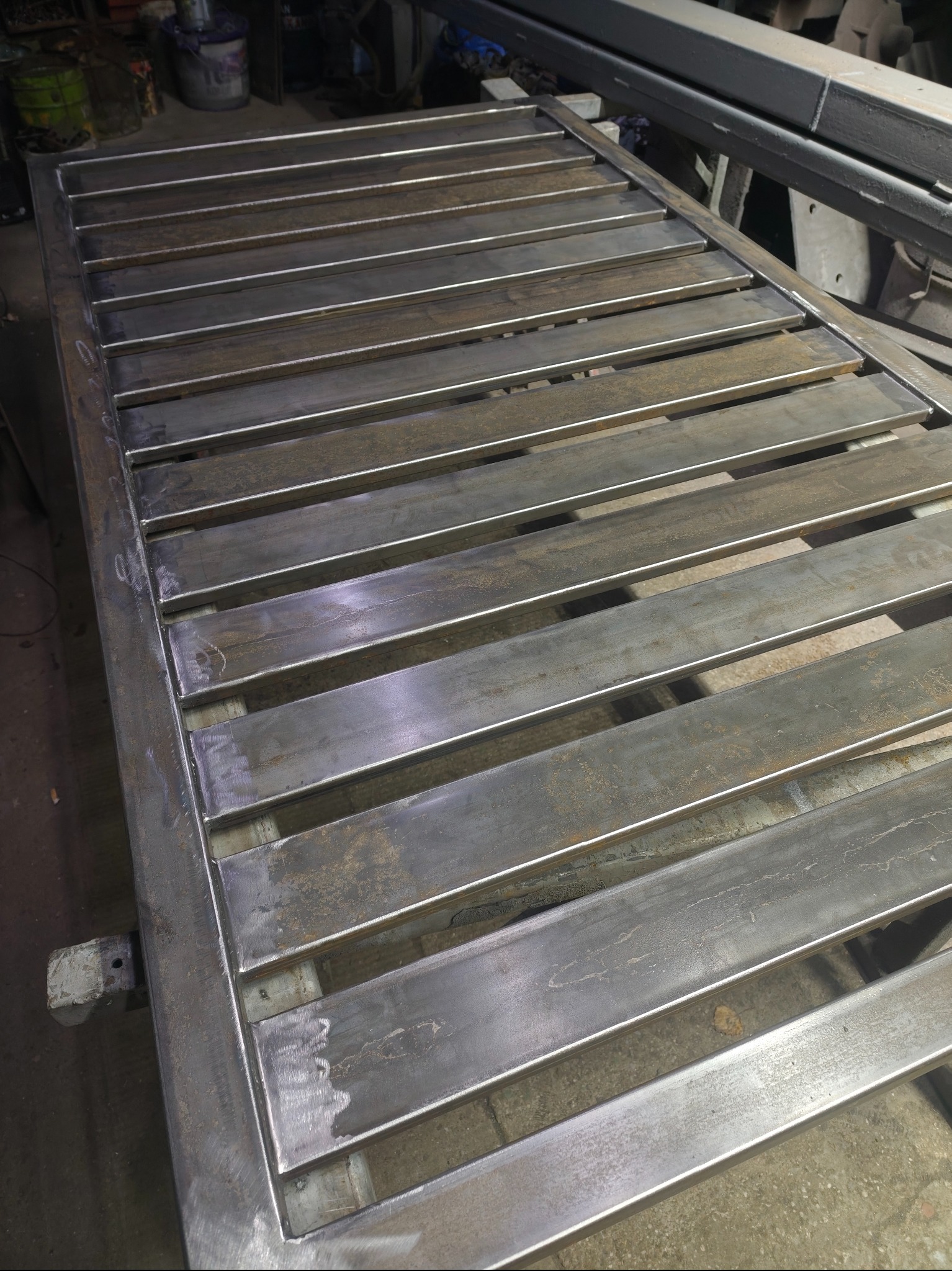 Surowa, metalowa konstrukcja bramy z poziomymi szczeblami, widoczne ślady spawania i obróbki, leżąca w warsztacie. Detal wykonania bramy przemysłowej.