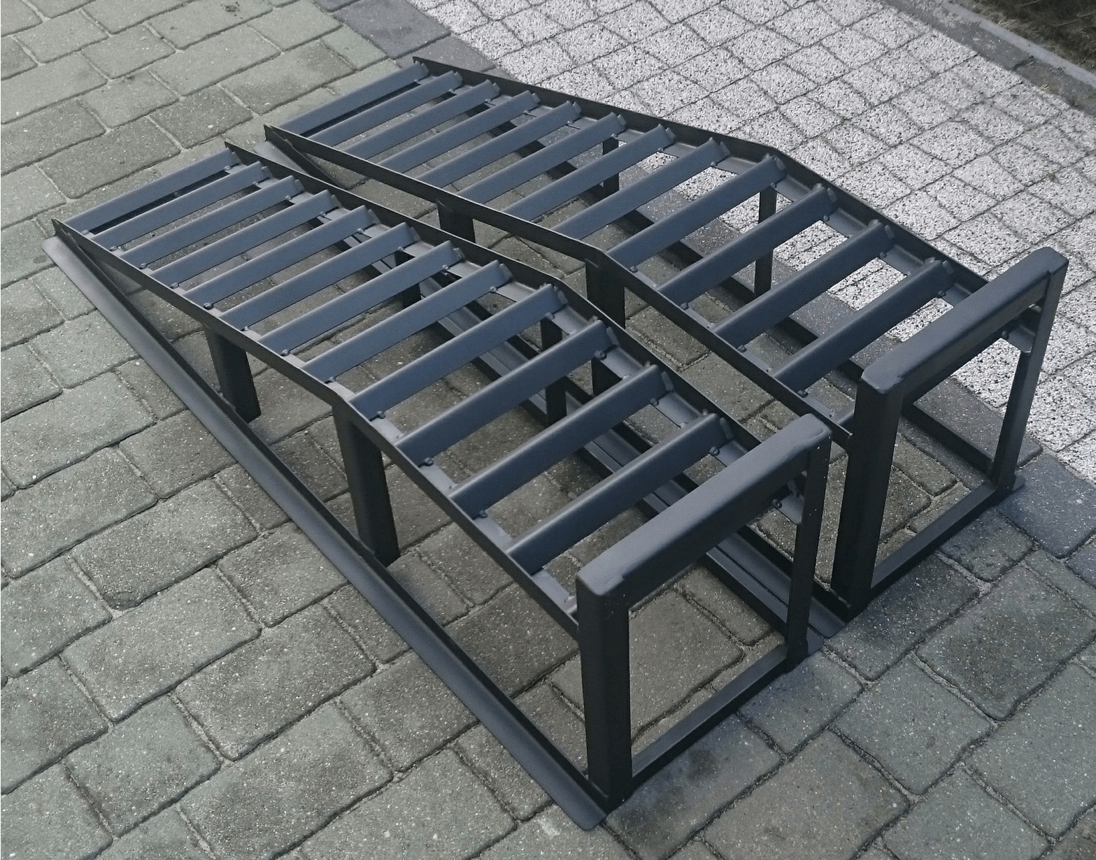 Dwie metalowe rampy najazdowe, pomalowane na szaro, ustawione na szarej kostce brukowej. Konstrukcja spawana z profili zamkniętych, widoczne spawy. Ujęcie z góry.