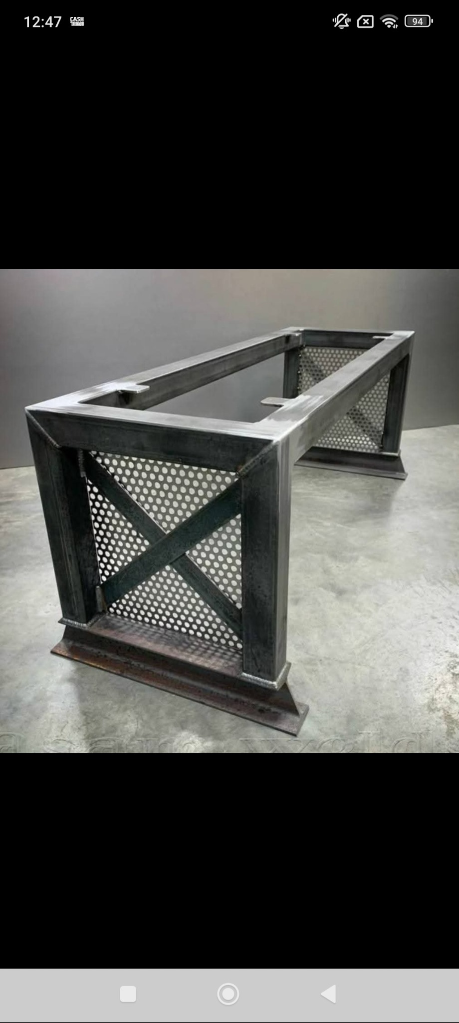 Metalowa konstrukcja stołu z perforowanymi panelami bocznymi w kształcie litery X, surowy industrialny styl, widoczne spawy, szara kolorystyka.