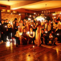 BedezetMoments - Grupa osób świętuje z zimnymi ogniami w dłoniach, w tle dekoracje i ciepłe światło, tworząc radosną atmosferę. Na pierwszym planie kieliszki i konfetti.