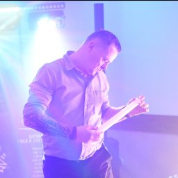 BedezetMoments - Mężczyzna z tatuażem na ramieniu, w koszuli, na scenie w fioletowym oświetleniu, trzymający biały przedmiot. W tle logo 'Kryss' i reflektory.