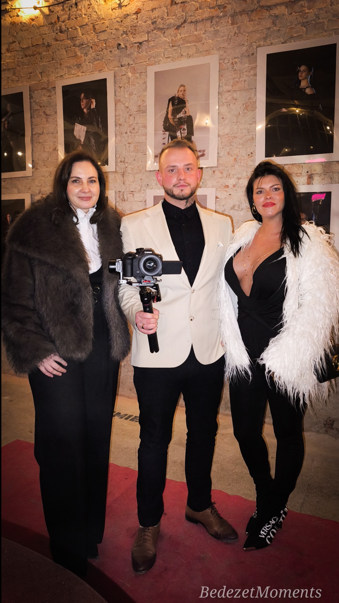 Kamerzysta z aparatem i dwie kobiety w eleganckich strojach pozują na tle ceglanej ściany z powieszonymi zdjęciami. Czerwony dywan dodaje szyku.