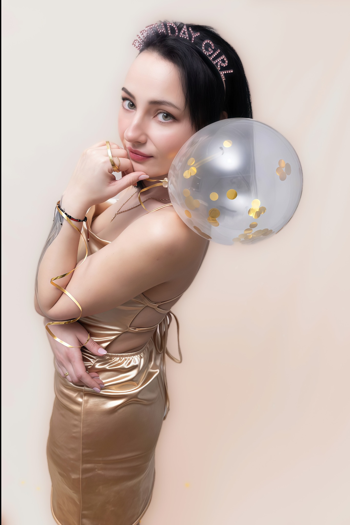 Portret kobiety w złotej sukience z balonem i opaską 'Birthday Girl', pozującej na jasnym tle. Uwagę przykuwają detale biżuterii i delikatny makijaż.