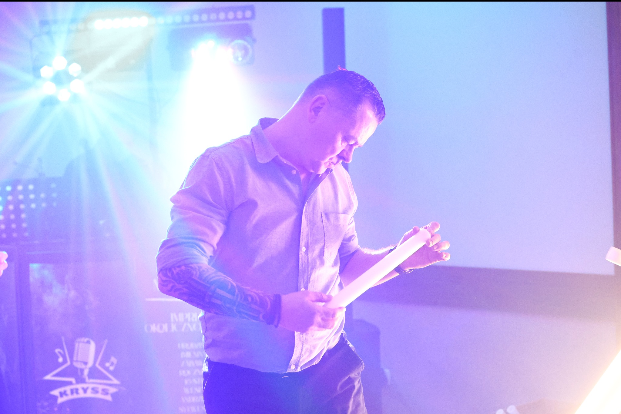 Mężczyzna z tatuażem na ramieniu, w koszuli, na scenie w fioletowym oświetleniu, trzymający biały przedmiot. W tle logo 'Kryss' i reflektory.